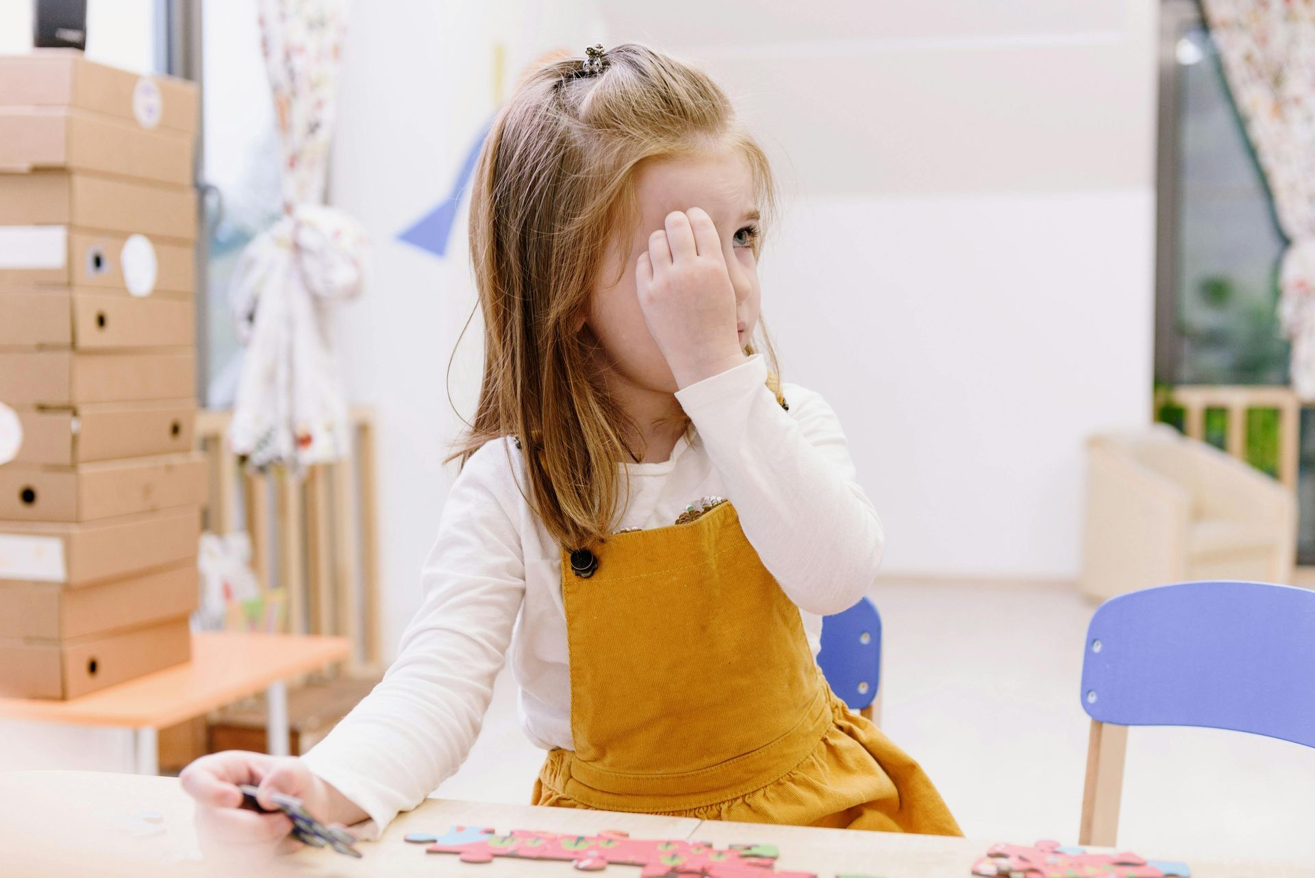 Child in a yellow dress covering one eye while sitting at a table in a bright classroom