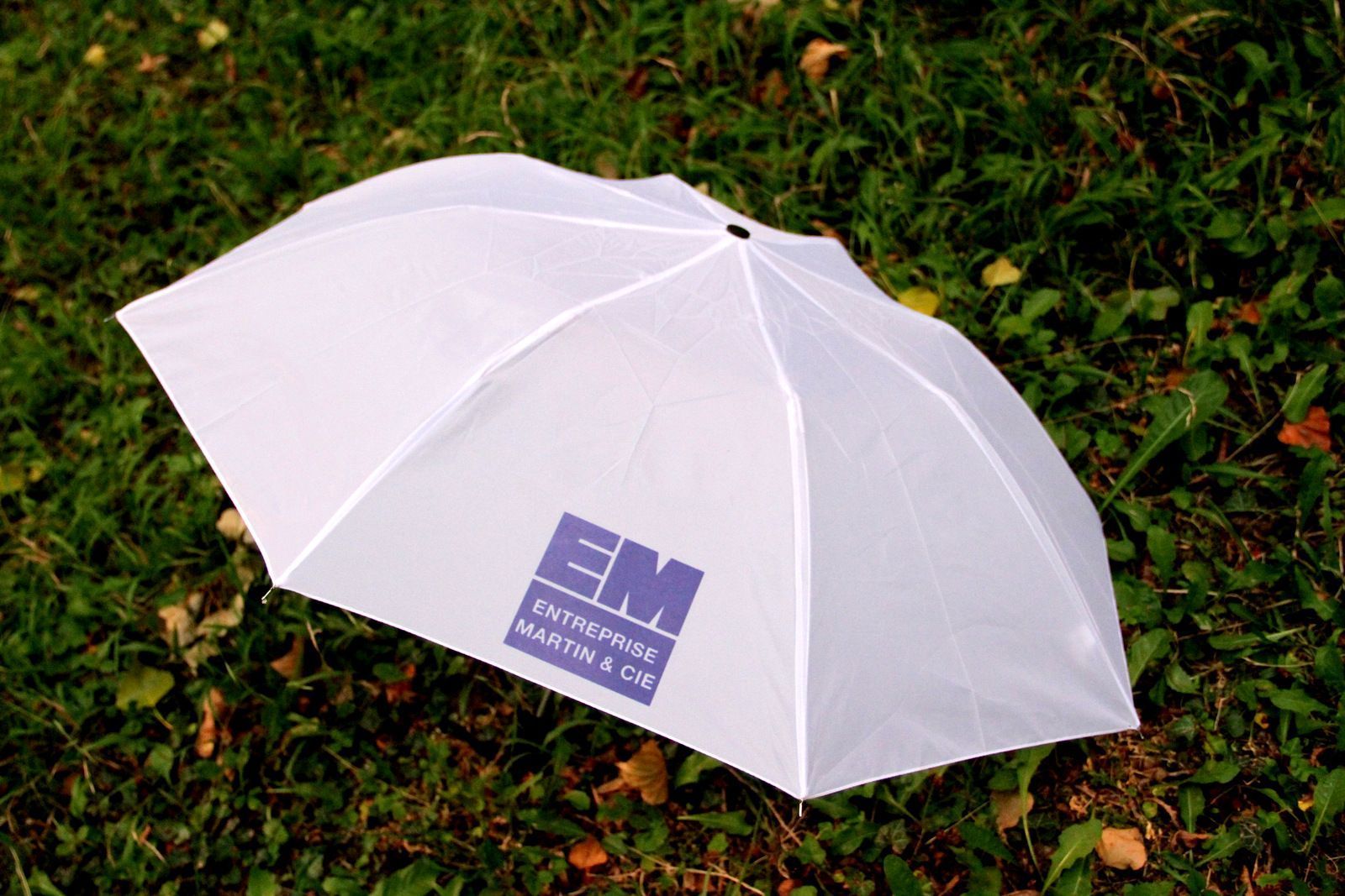 Parapluie blanc posé sur l'herbe verte, avec un logo bleu sur la toile