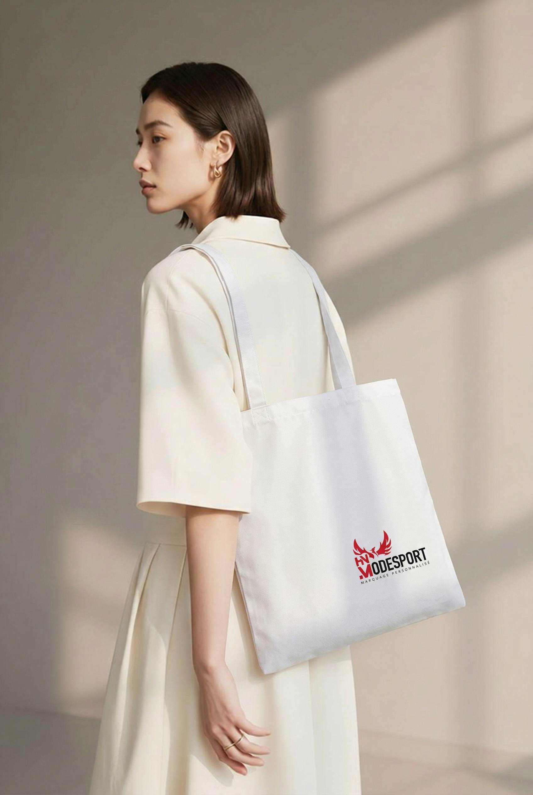 Une personne en robe crème portant un sac cabas blanc, adossée à un mur beige clair.