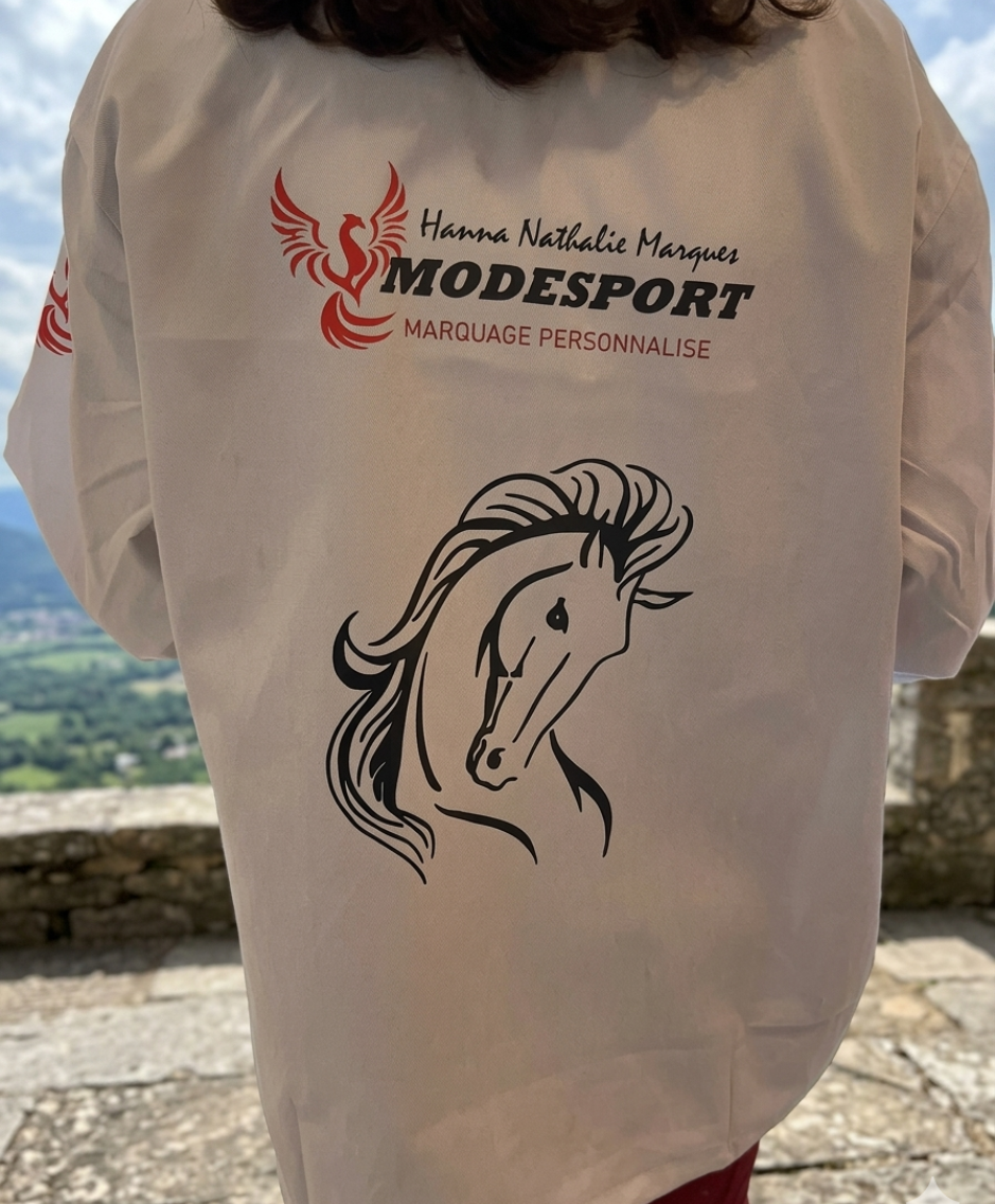 Une personne portant un t-shirt beige avec l'inscription « MODESPORT » et un dessin de cheval, se tient à l'extérieur, devant un paysage de vallée.