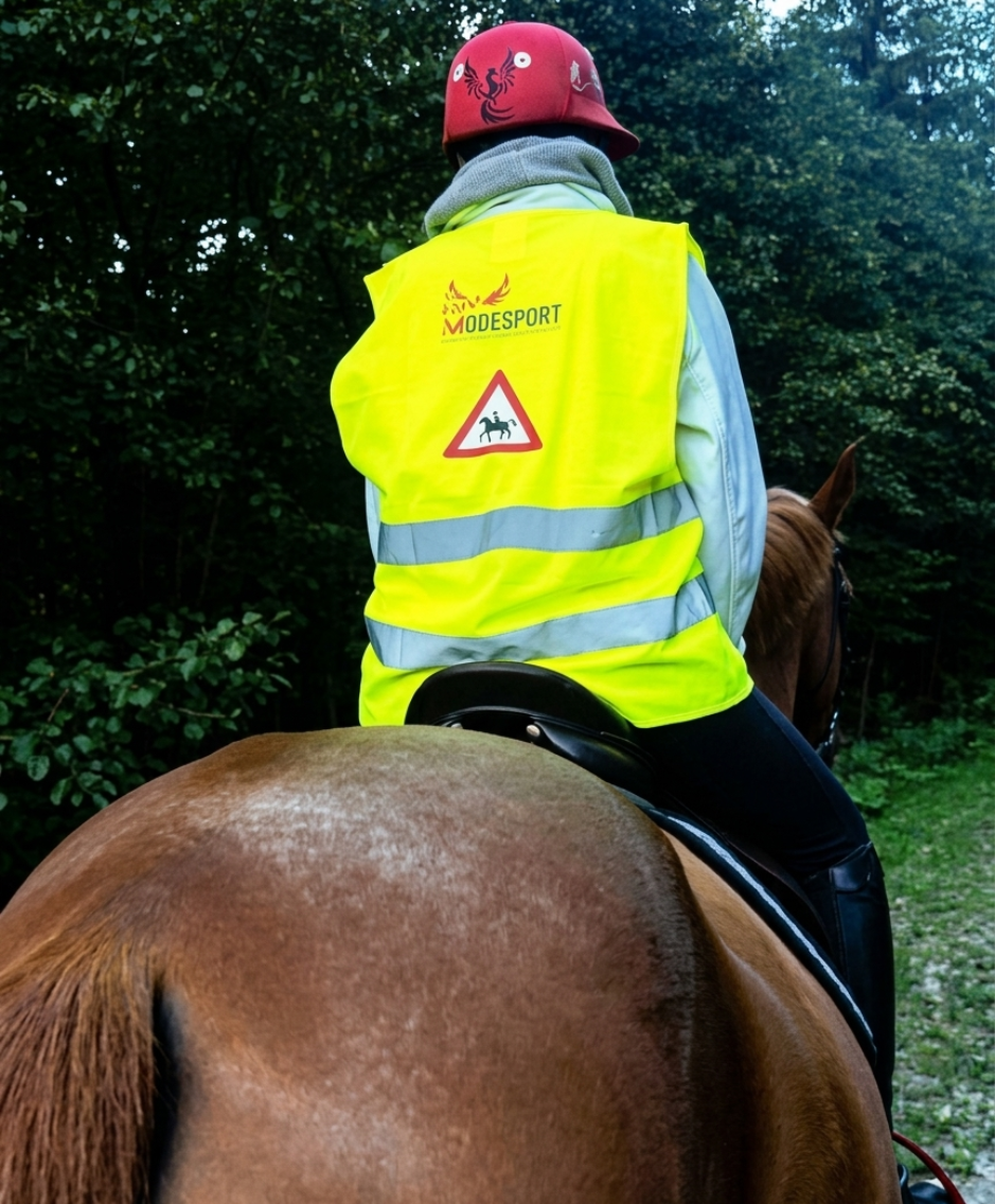 Une personne portant un casque rouge et un gilet de sécurité jaune monte à cheval sur un sentier boisé.