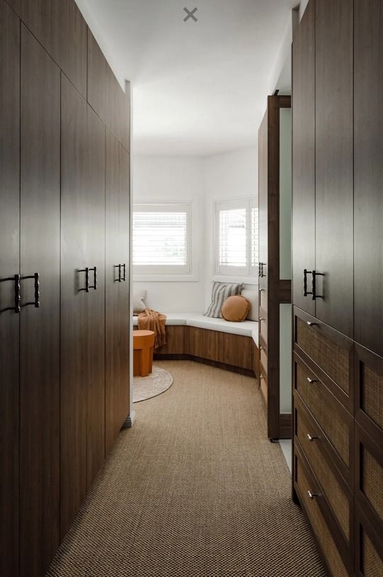 A narrow walk-in closet with dark wood cabinetry on both sides leading to a bright, cozy window seat at the end. — PKJ Design in Unanderra, NSW 