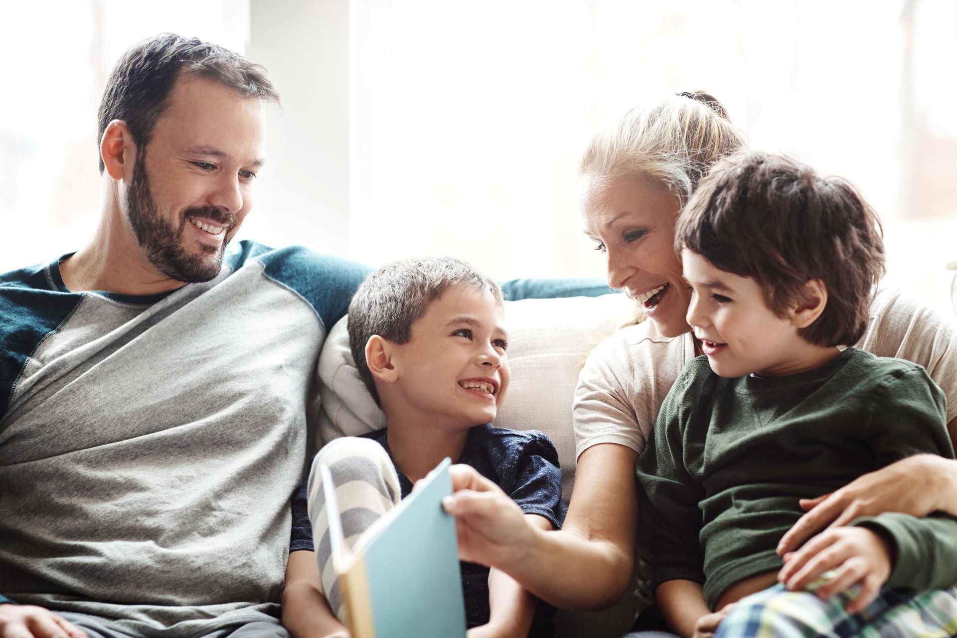 A smiling family of four sitting on a couch, reading a book together in a bright living room.