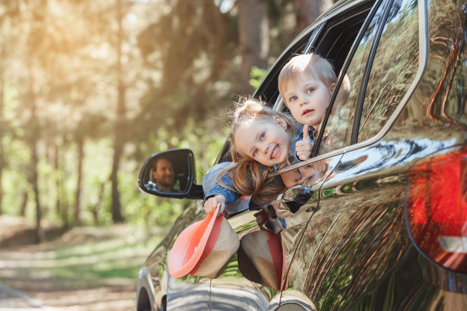 Two children leaning out of a car window, smiling in a sunny wooded area