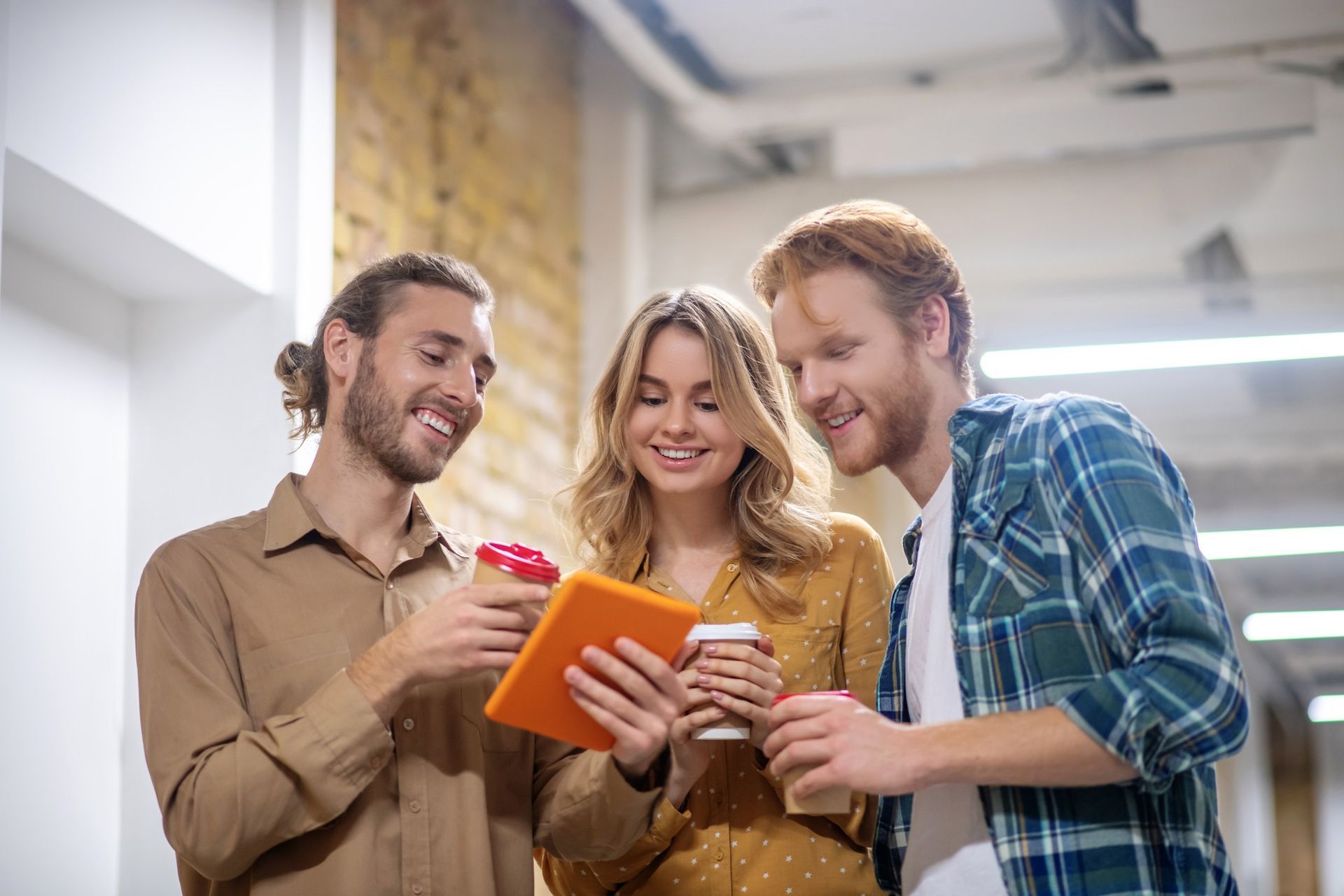 Three coworkers smiling and sharing notes on a tablet in a bright office hallway