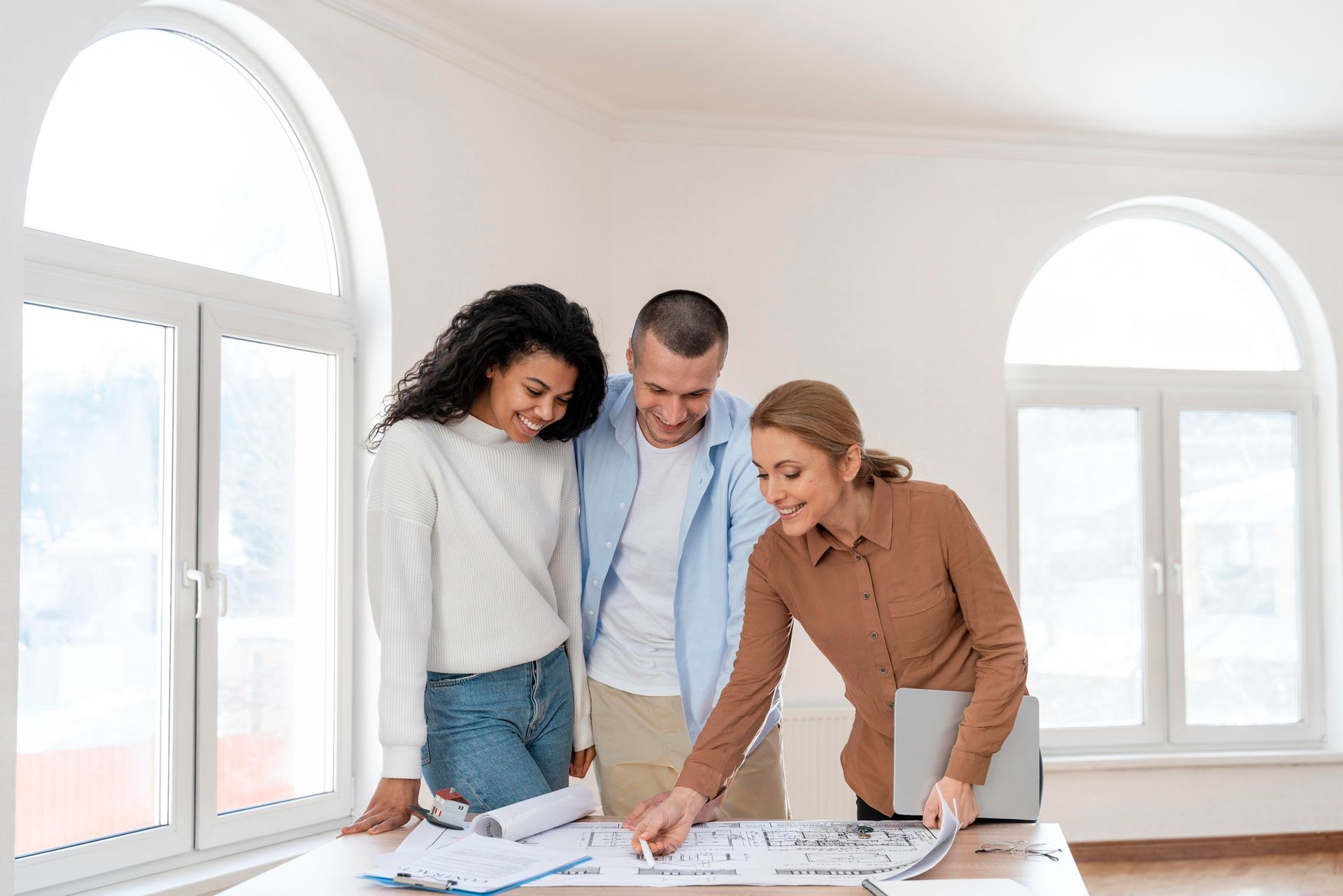 Three people reviewing architectural plans on a table in a bright room with large arched windows