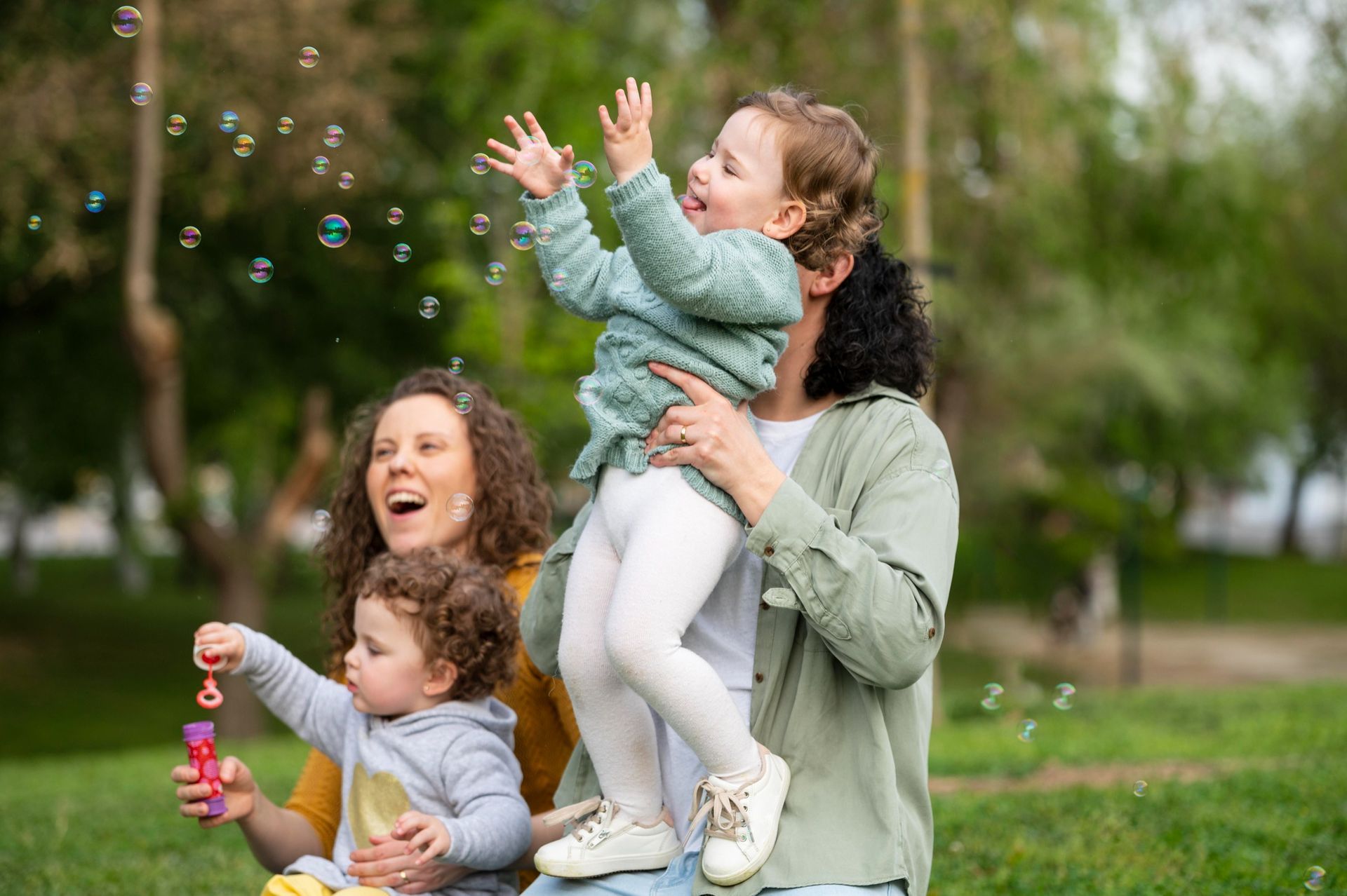 Family blowing bubbles in a park, with a child lifted in the air and everyone smiling.