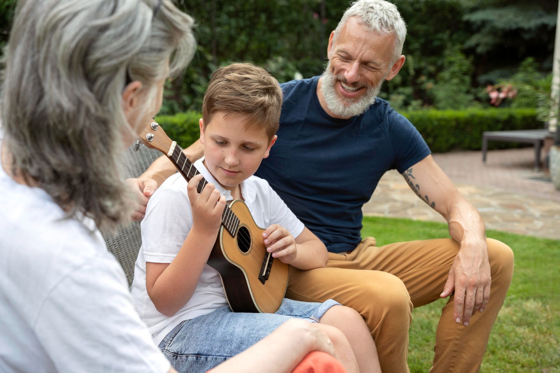 Two people listening as a boy plays a small guitar in a park