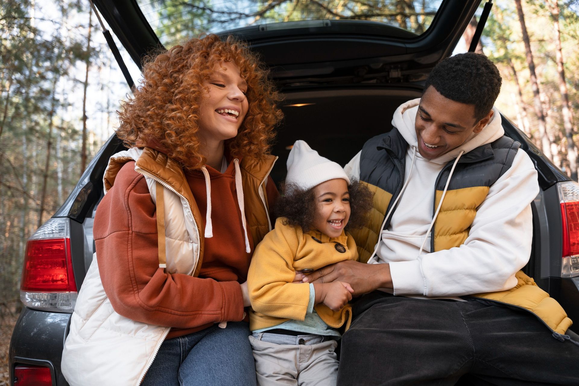 Family smiling by open car trunk outdoors, with a child sitting between two adults.