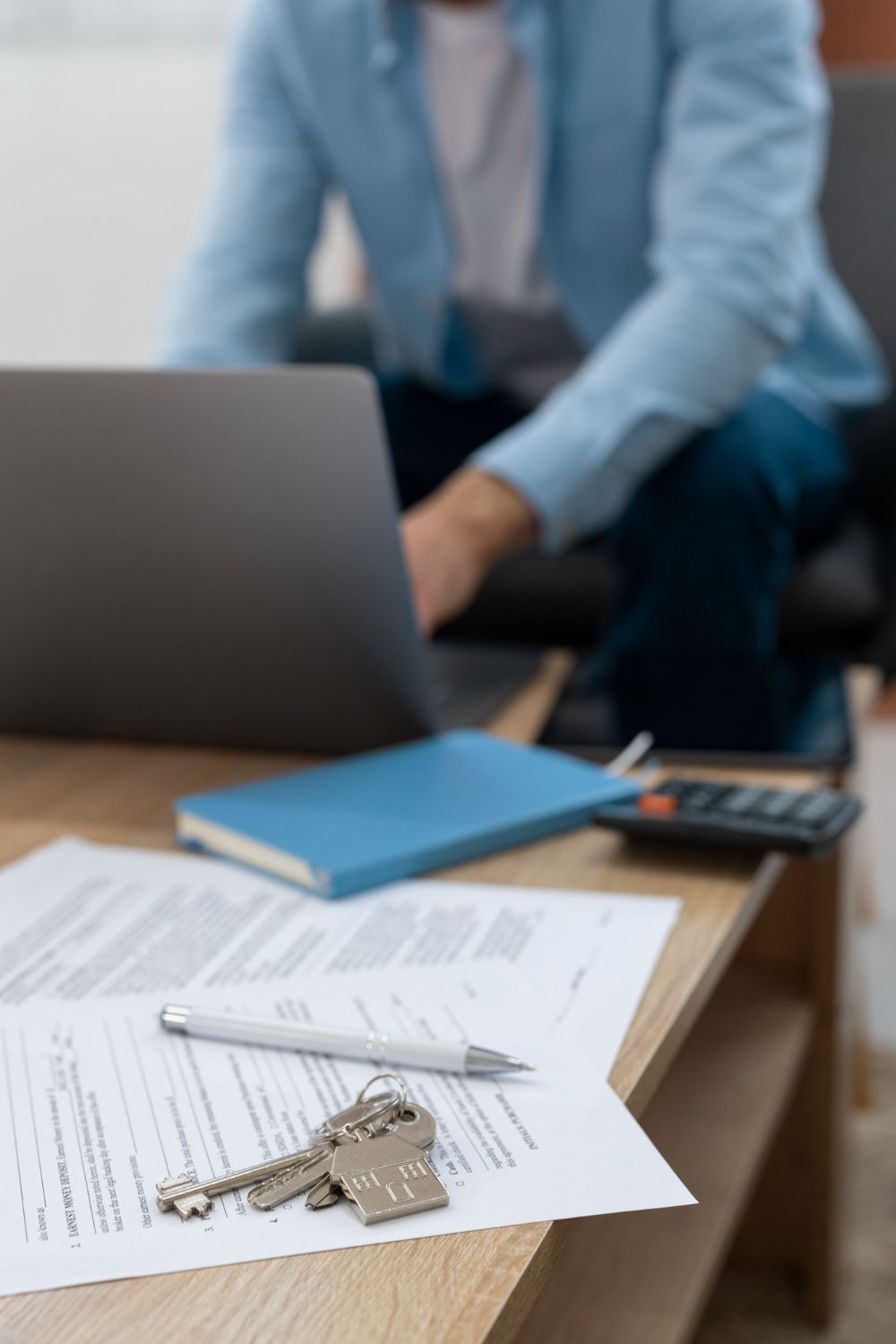 Office desk with laptop, papers, keys, notebook, calculator, and person working in the background