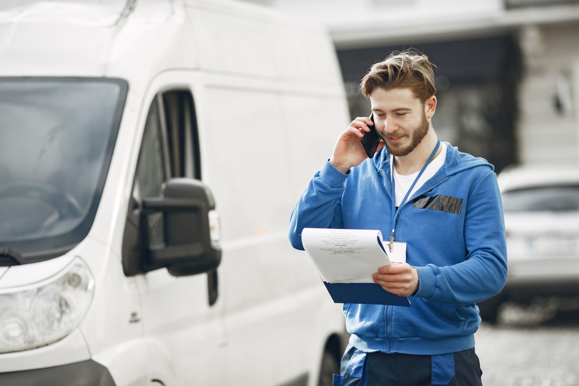 Courier in blue jacket reading a clipboard beside a white delivery van while talking on a phone