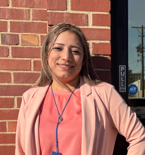 Smiling person in a coral blazer standing against a brick wall by a glass door