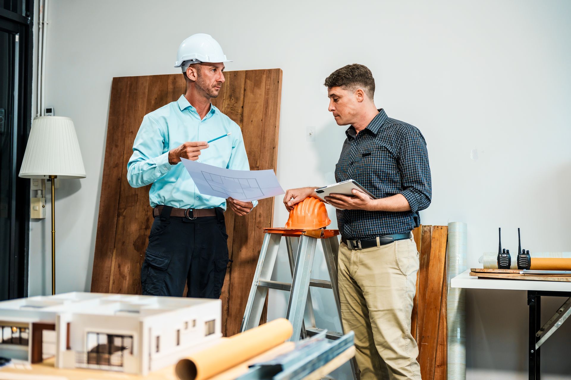 Two workers in hard hats review blueprints beside a model house in a workshop