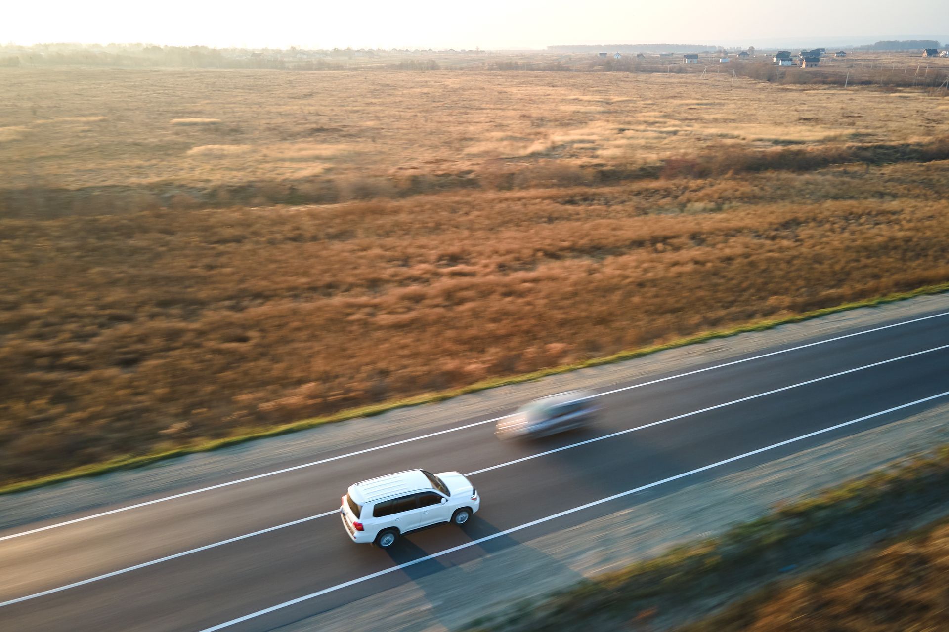 White SUV drives on a highway beside a dry, open field at sunset