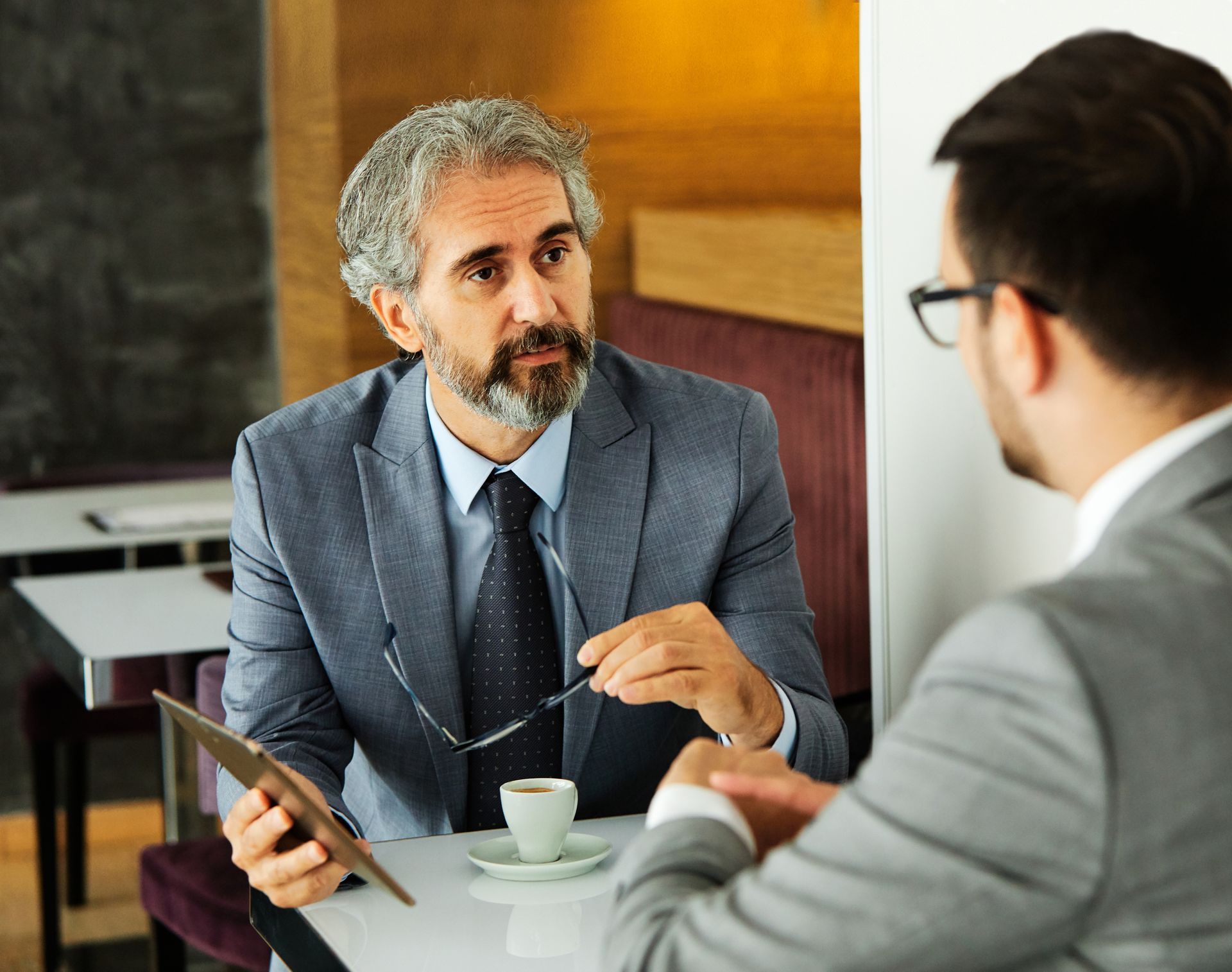 Two men in suits discuss over coffee at a table, one holding a tablet and gesturing with a pen.