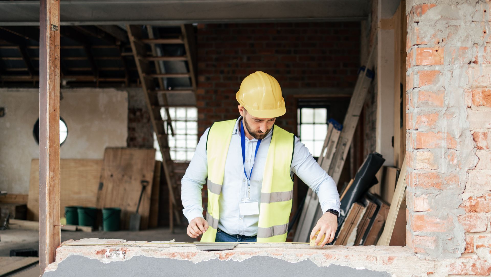 Construction worker in yellow hard hat and reflective vest measuring materials at a building site.