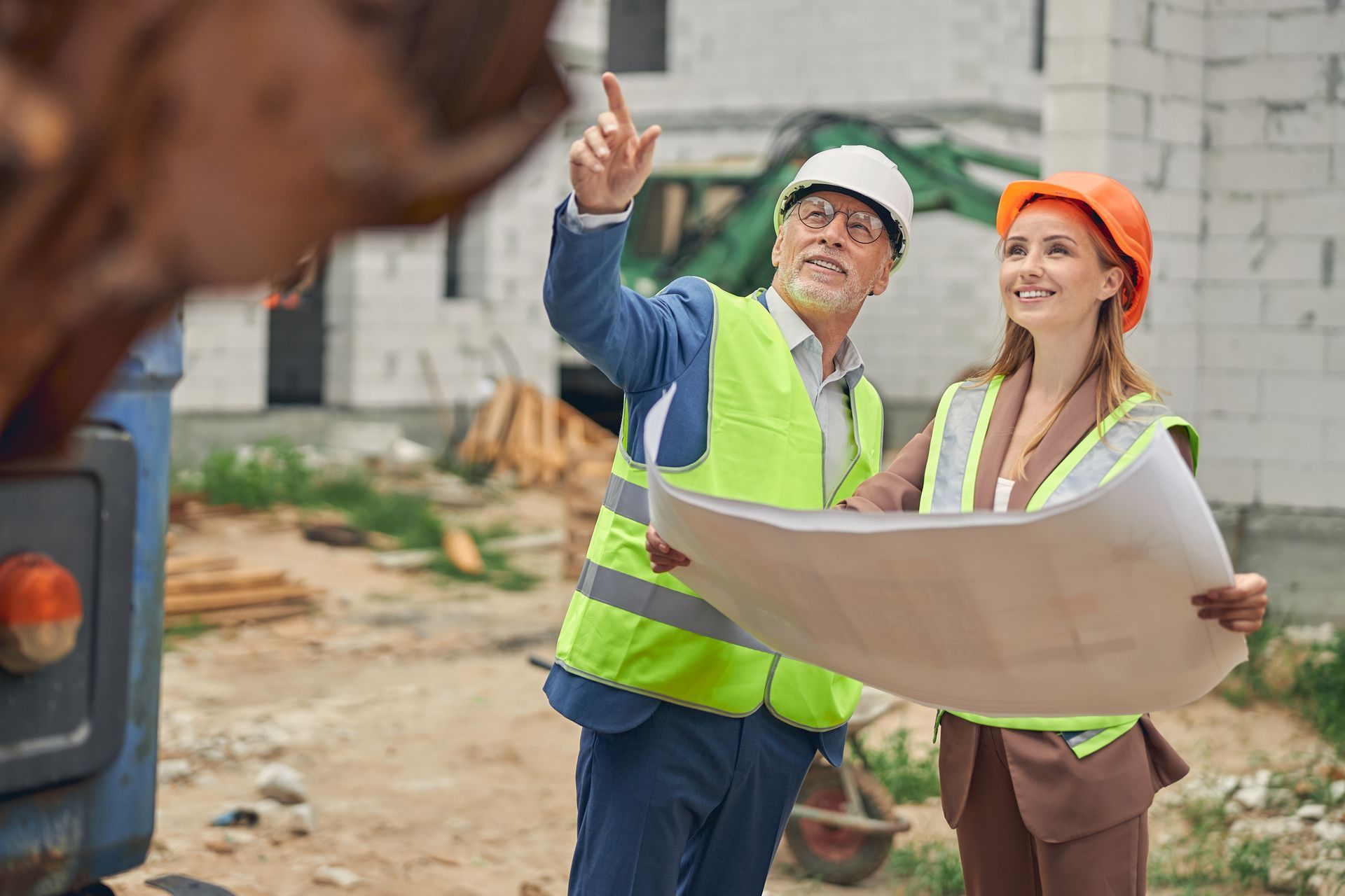 Two construction workers in hard hats review blueprints at a building site.