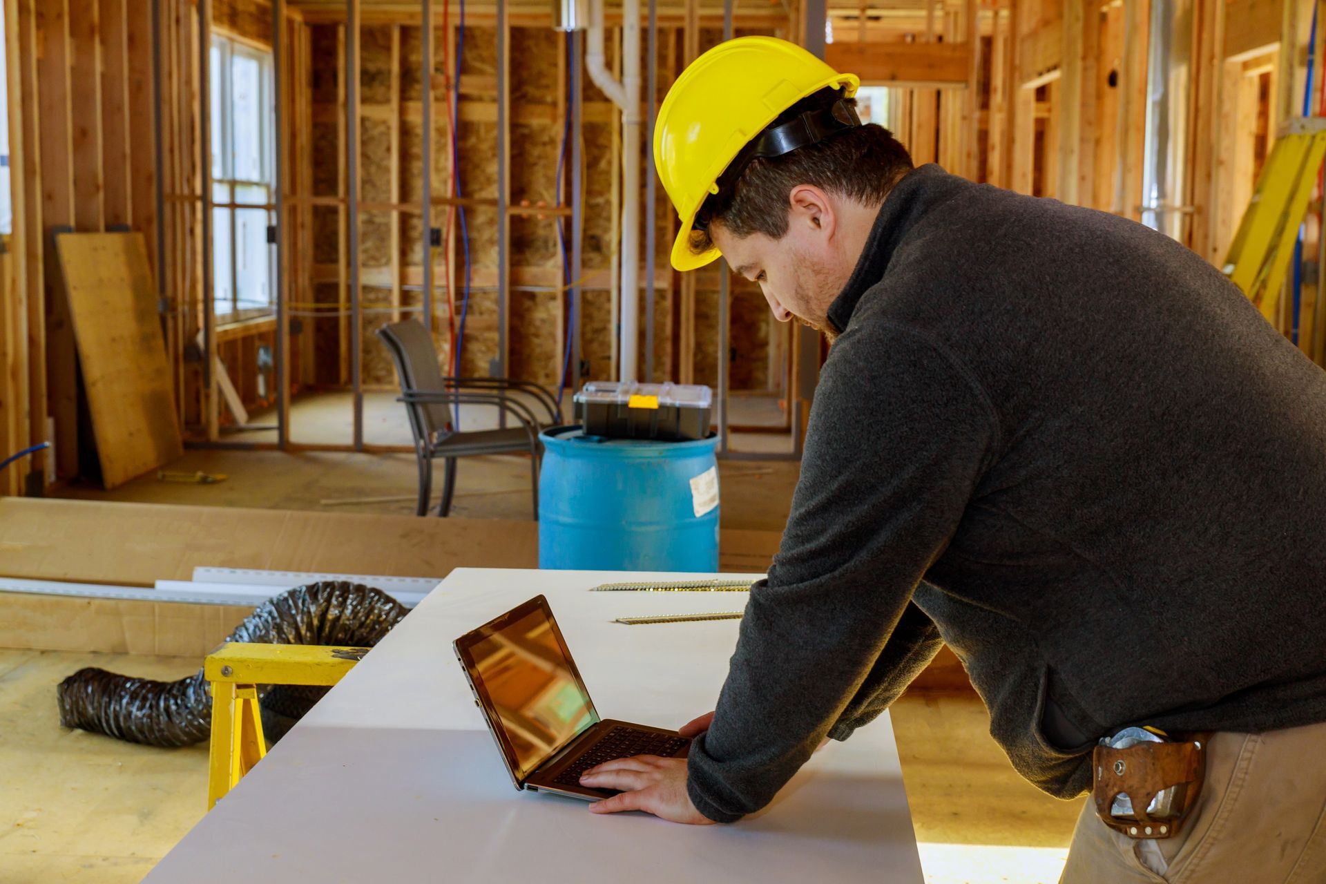 Construction worker in a hard hat using a laptop at a partially built house interior.