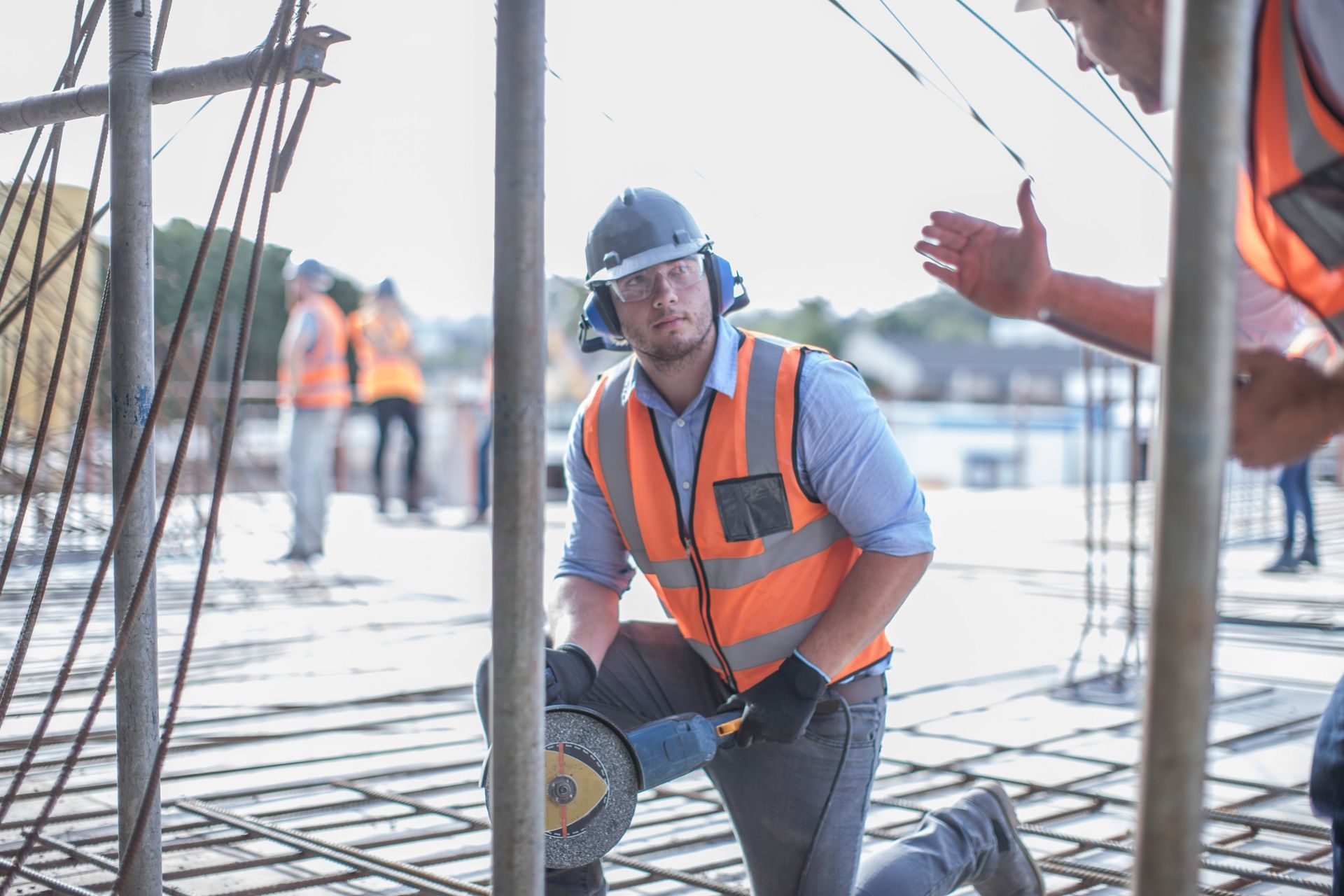 Construction worker kneeling with a power saw on a rebar floor slab, wearing helmet and orange safety vest