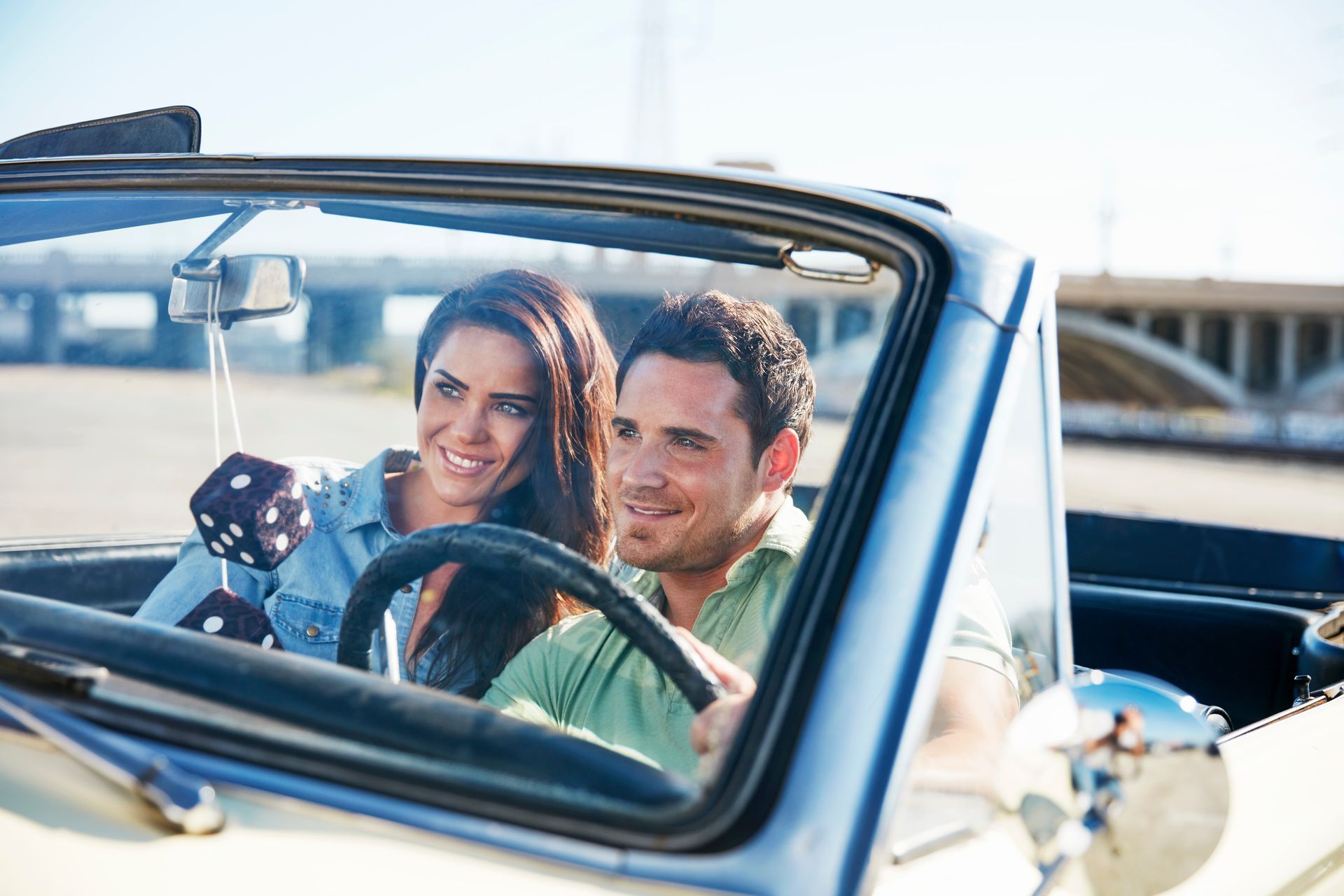 Couple smiling in a blue convertible, driving near the waterfront with dice hanging from the mirror