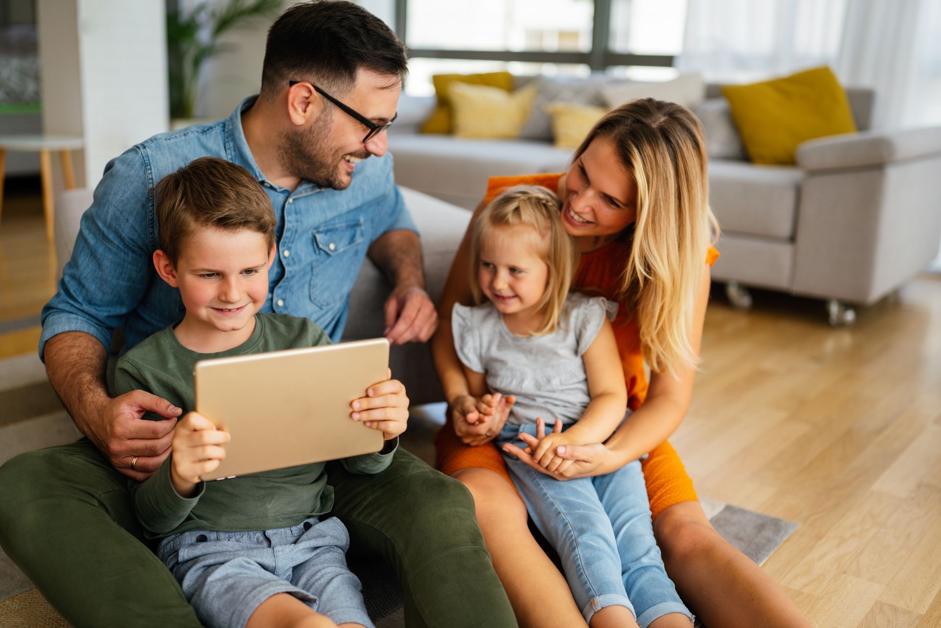 Family sitting on the floor together, looking at a tablet in a bright living room.