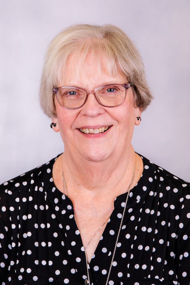 Smiling woman with glasses and short blonde hair, wearing a black polka-dot blouse against a light background