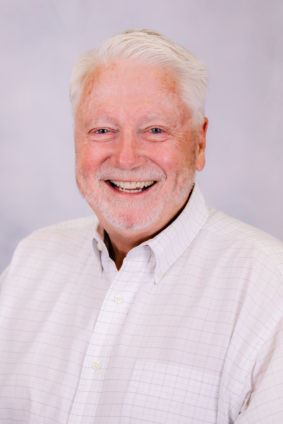 Smiling man with short white hair and beard in a light button-up shirt against a plain background