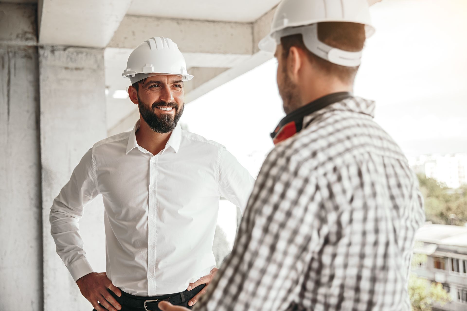 Two construction workers in hard hats talk on a building site, one smiling with hands on hips.