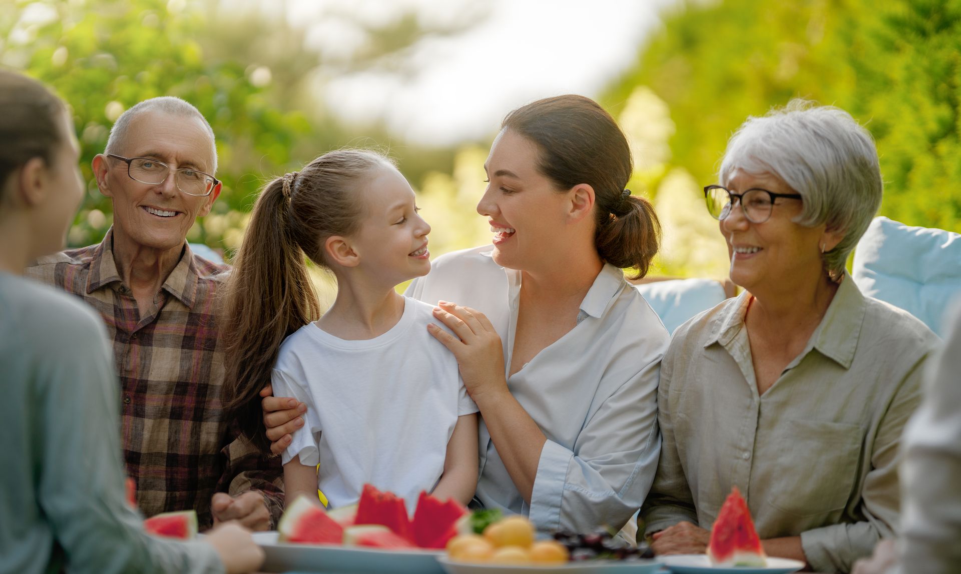 People sharing drinks and watermelon outdoors at a picnic table, smiling in warm sunlight