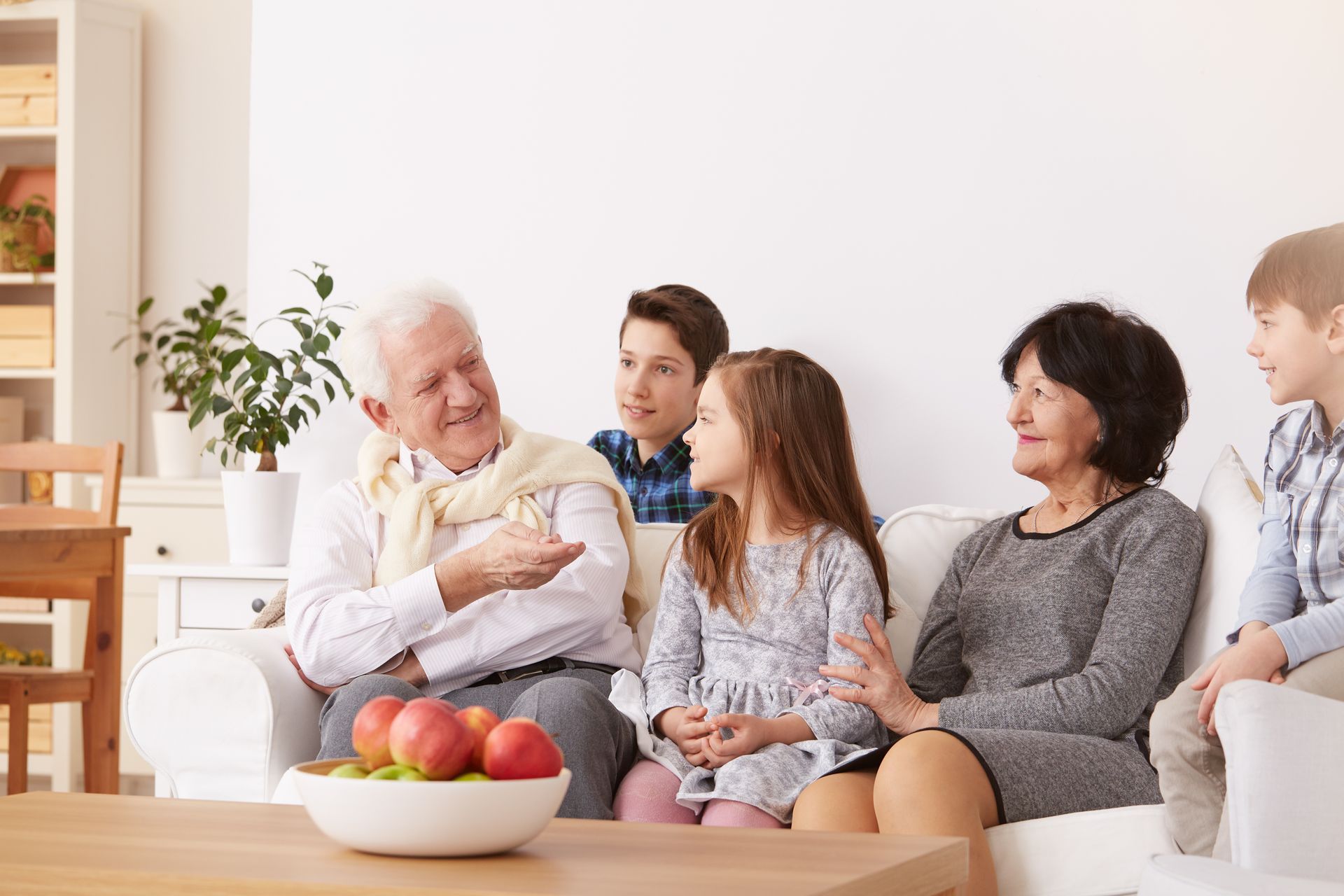Family sitting together on a couch in a bright living room, chatting and smiling over a bowl of fruit.