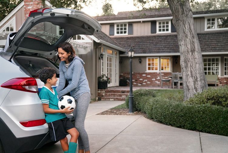 Person loading a child with a soccer ball into the open trunk of a white SUV in a residential driveway