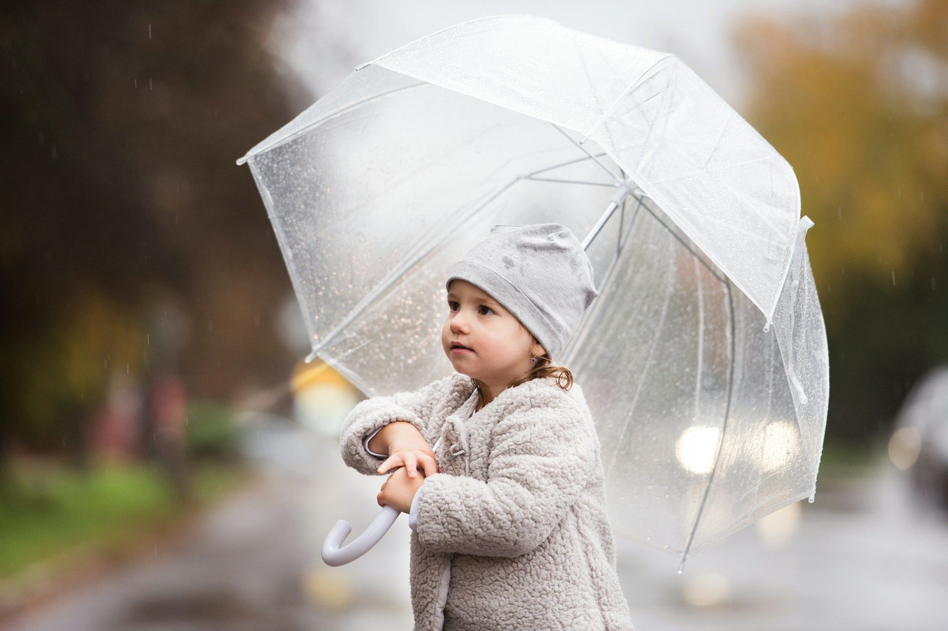 Child holding a transparent umbrella on a rainy sidewalk, wearing a light gray coat and hat.