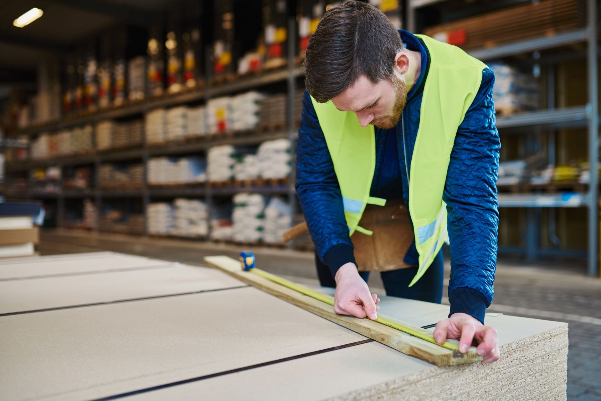 Worker in a warehouse measuring a countertop with a tape measure, wearing a yellow safety vest.