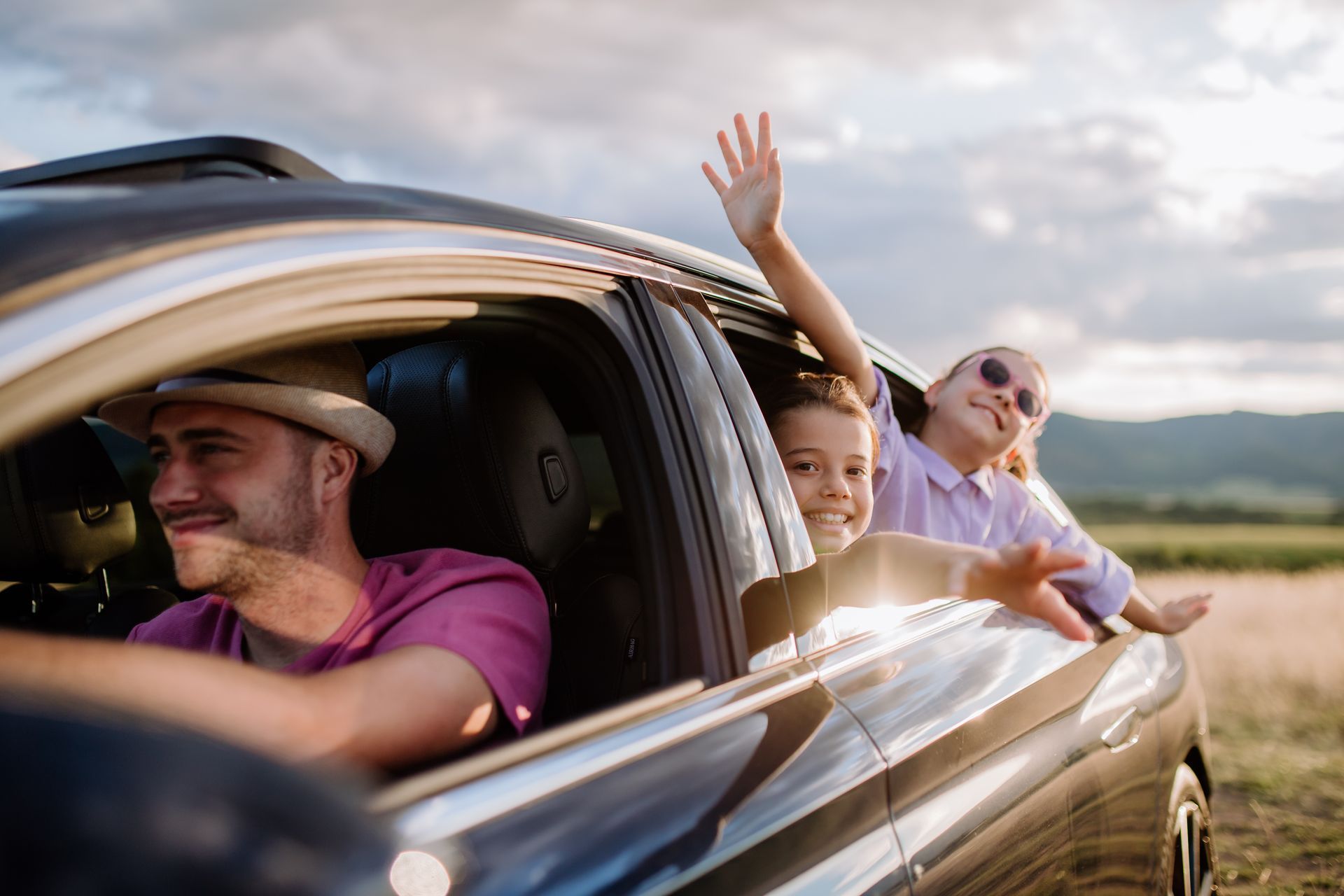 Friends leaning out of a car window on a sunny country road, smiling and waving