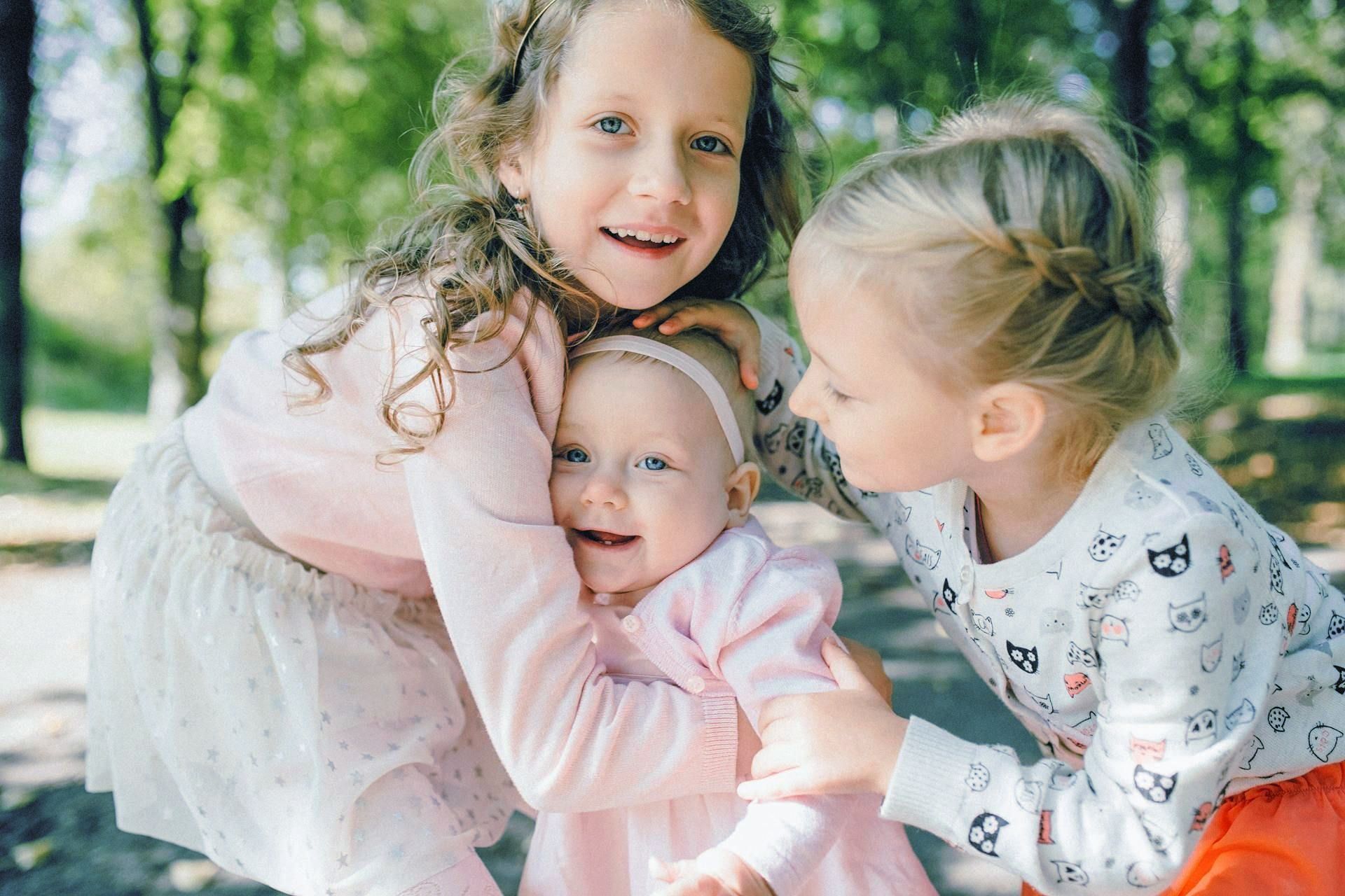 Three smiling children hugging outdoors, wearing pastel clothes.