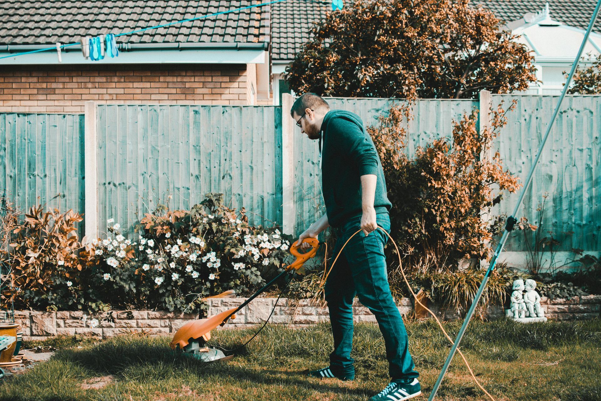 Person watering a garden with a yellow hose beside flowers and a fence.