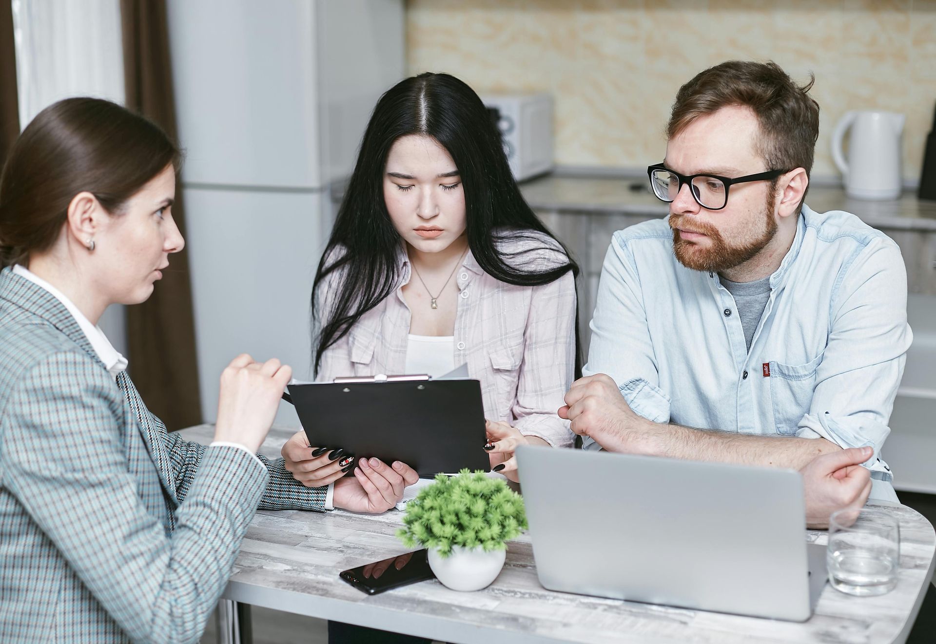 Three coworkers discuss a tablet and laptop at a table in a bright office.