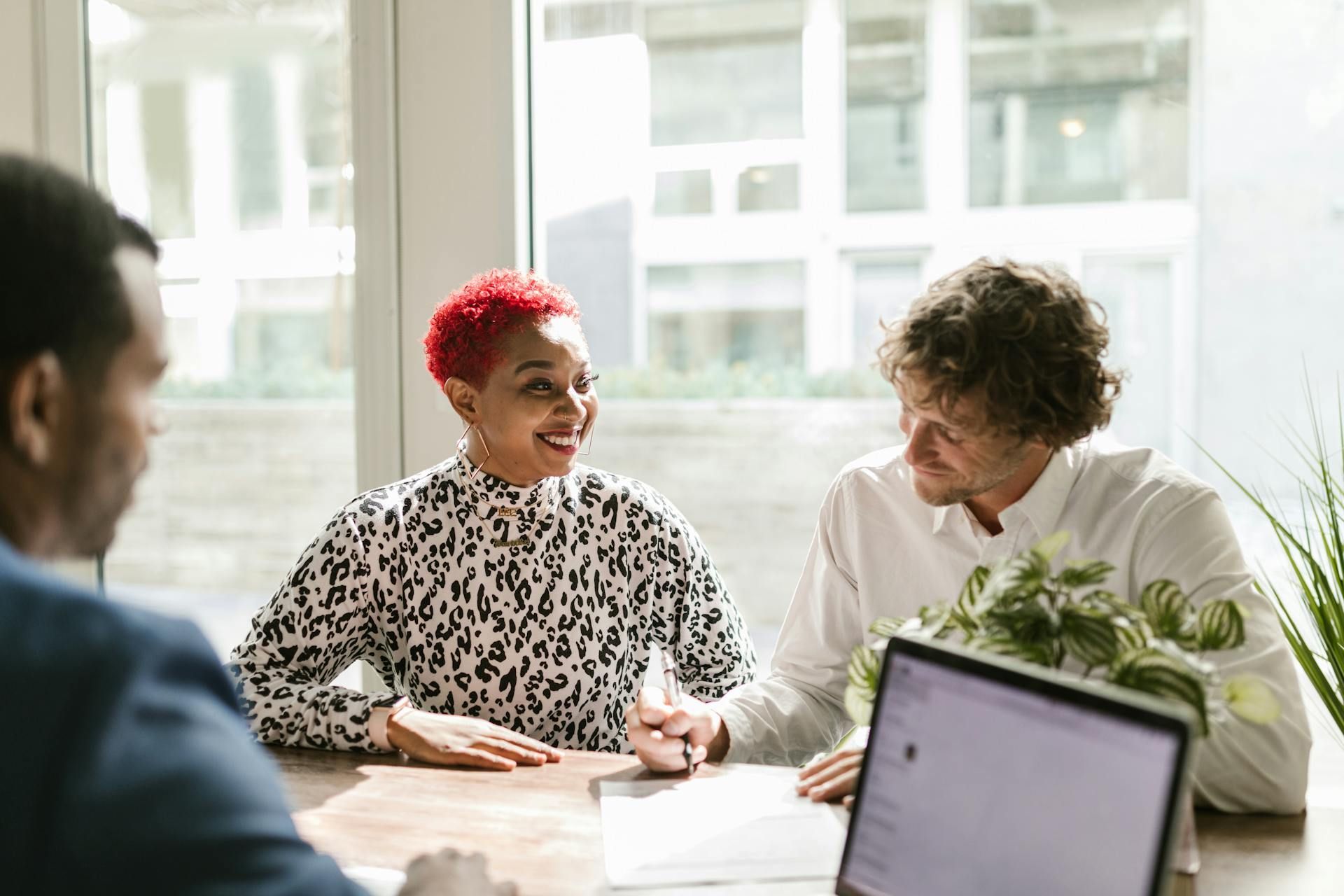 Three people in a bright office meeting, smiling and talking at a table with a laptop and plant.