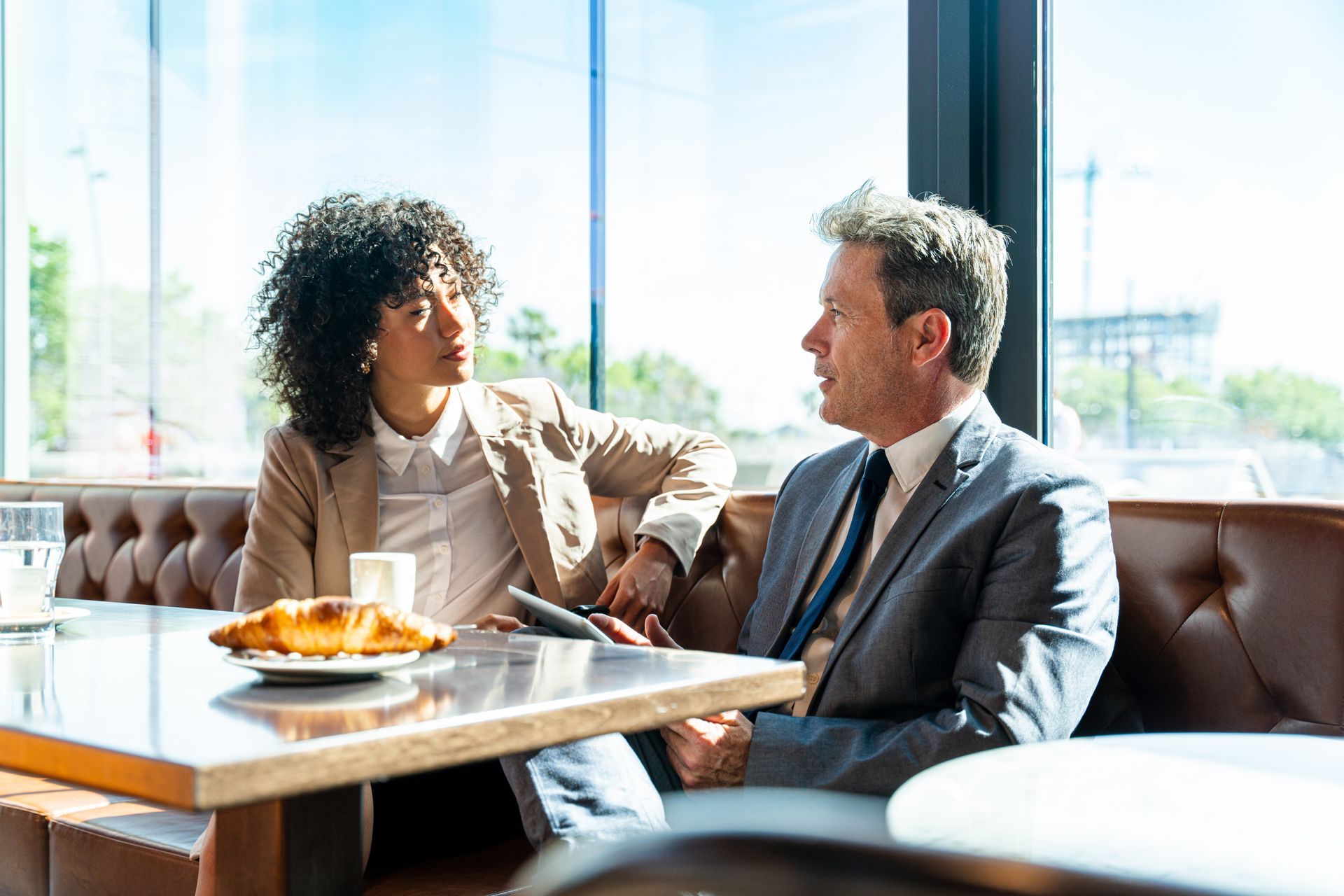 Two coworkers in business attire talking at a table in a bright café with a plate of food nearby
