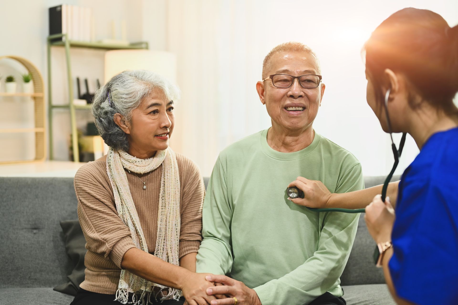 Healthcare worker checking a seated patient’s heartbeat while an older couple smiles on a couch