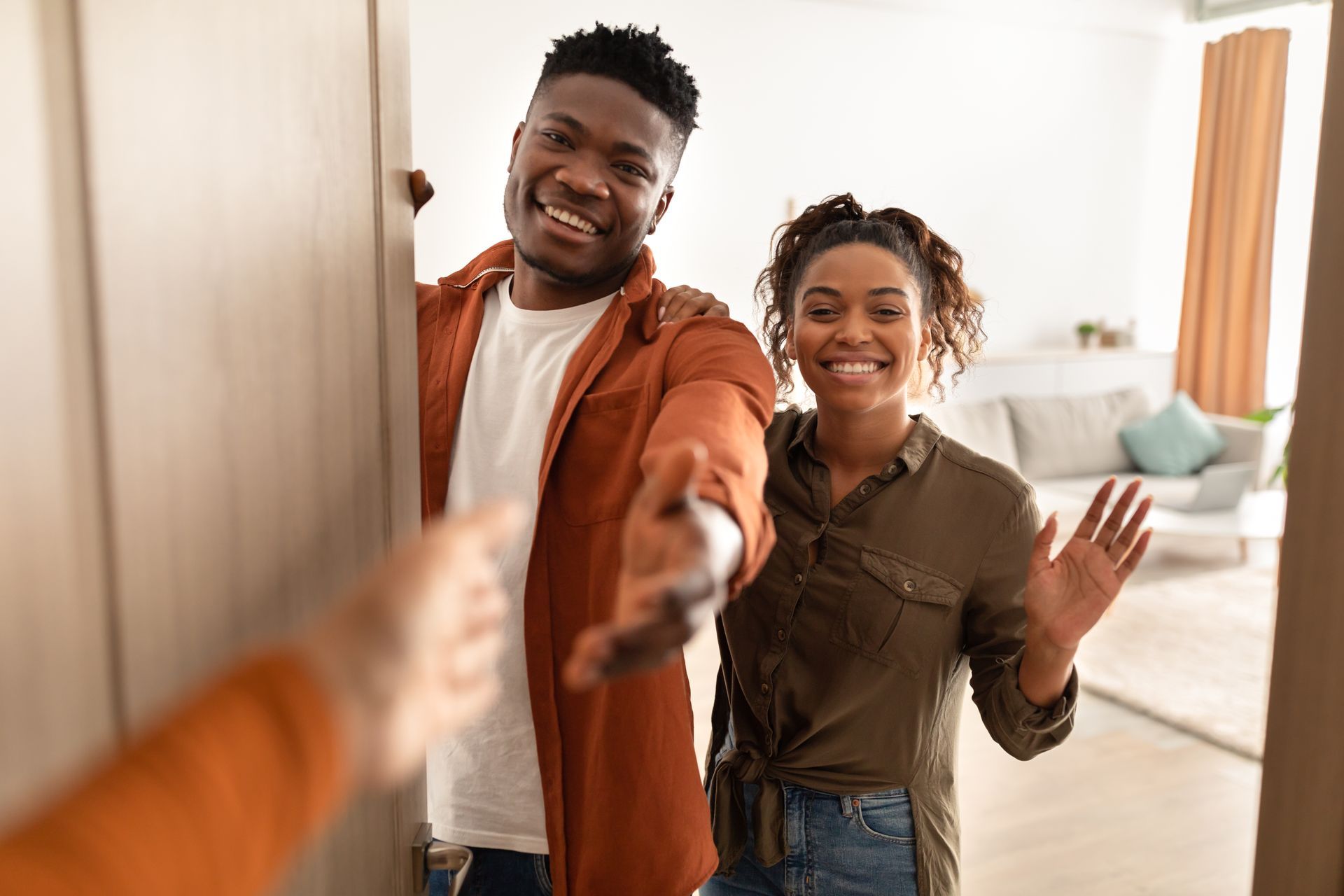 Smiling couple opening the front door, one reaching in to greet them in a bright living room