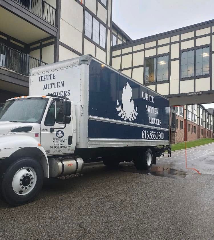 White moving truck parked by a building on a wet street, with a bridge walkway overhead.