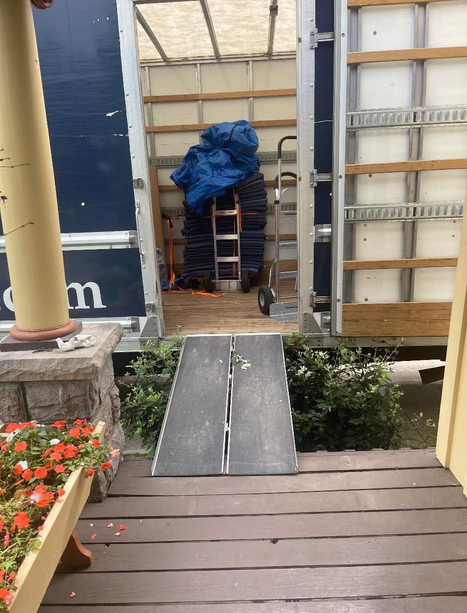 Wooden porch with a narrow ramp leading to a yard, framed by flowers and a ladder against a wall.