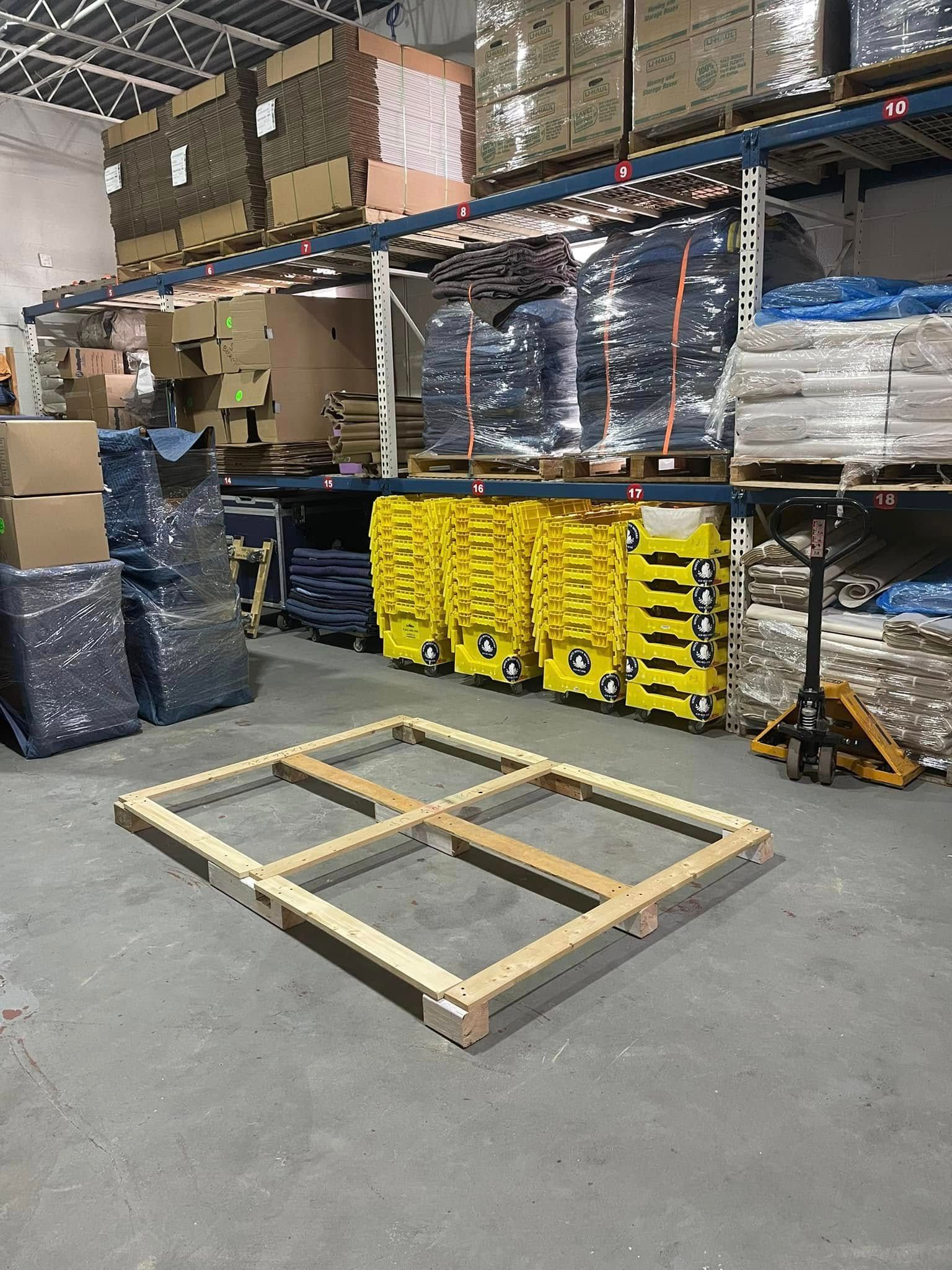Warehouse with a wooden pallet frame on the floor, shelves of boxed goods, and stacked yellow carts in the background