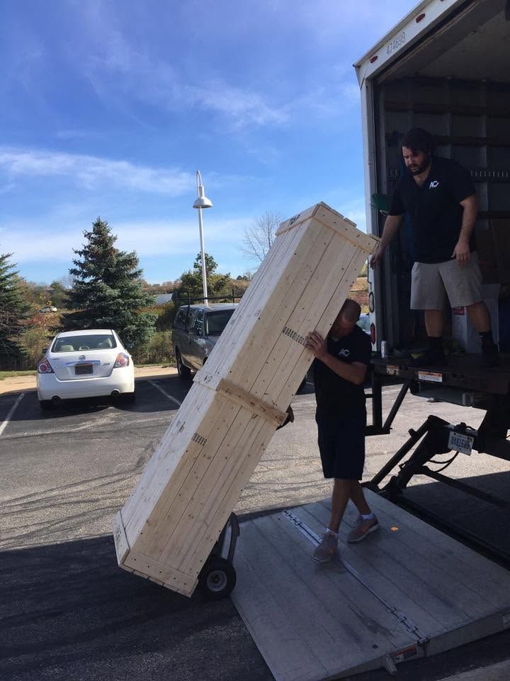 Two people unloading a large stacked package from a truck ramp outdoors.