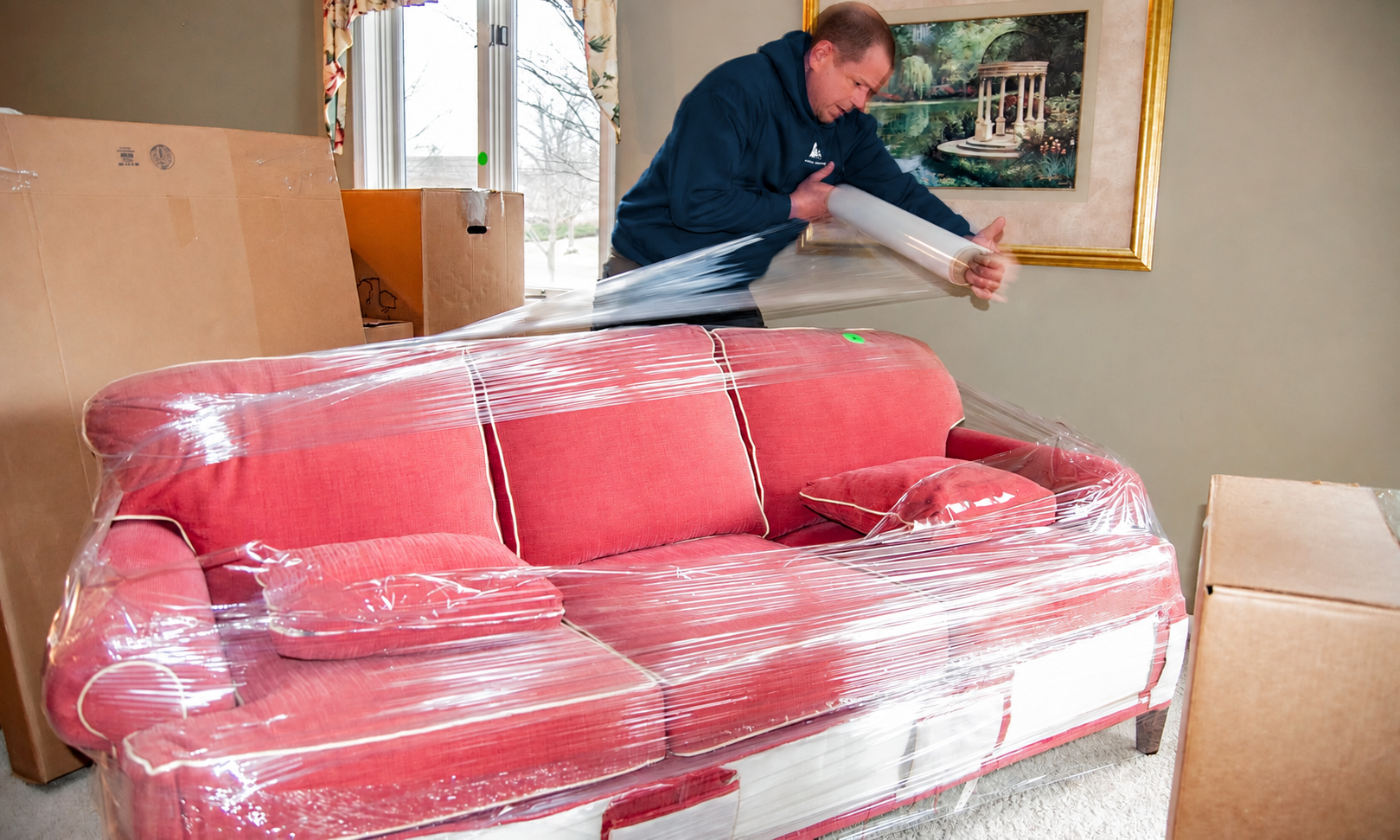 Man wrapping a red sofa in clear plastic in a moving room