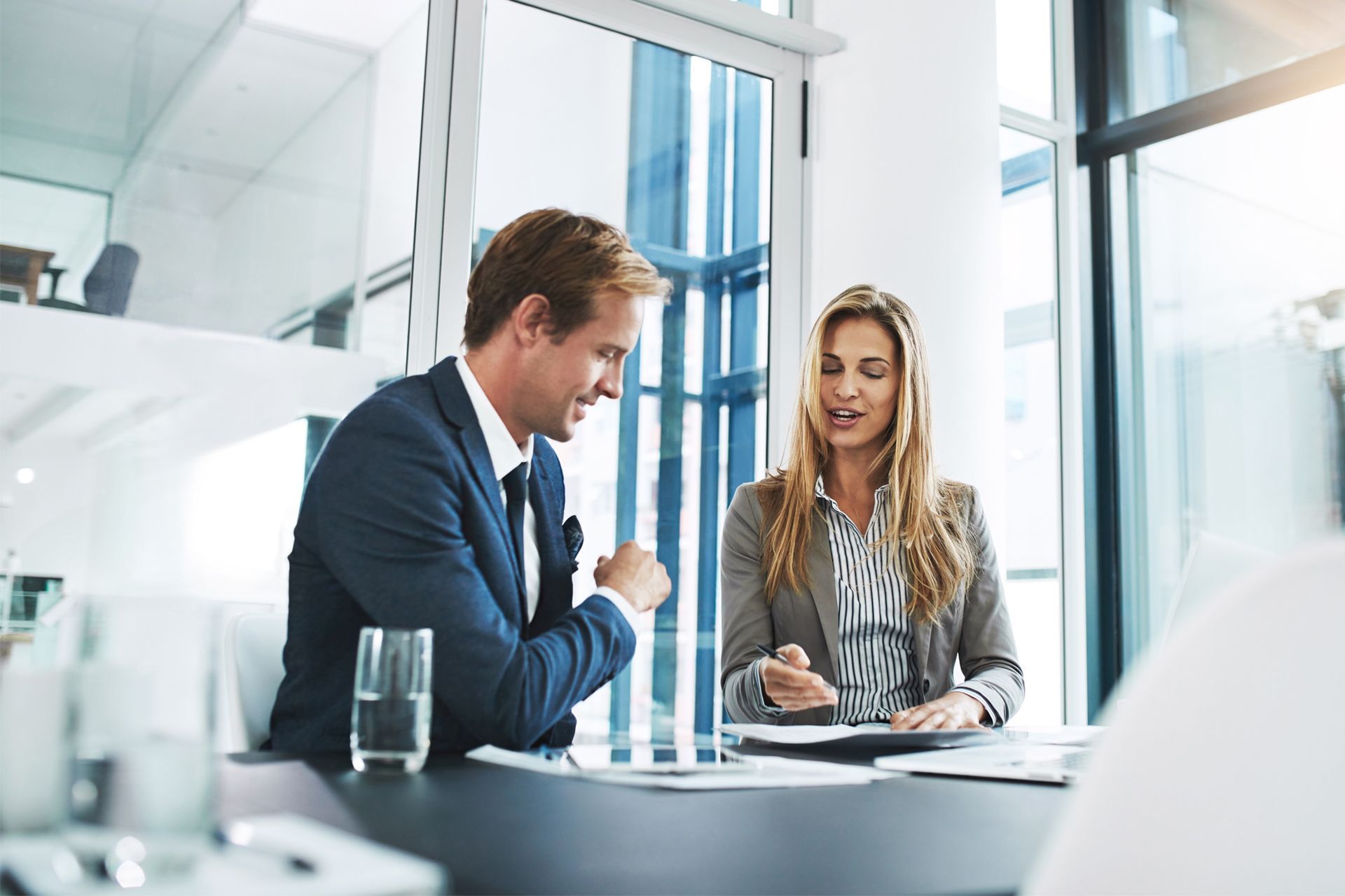 Man and woman in suits review documents at a table in a modern office.