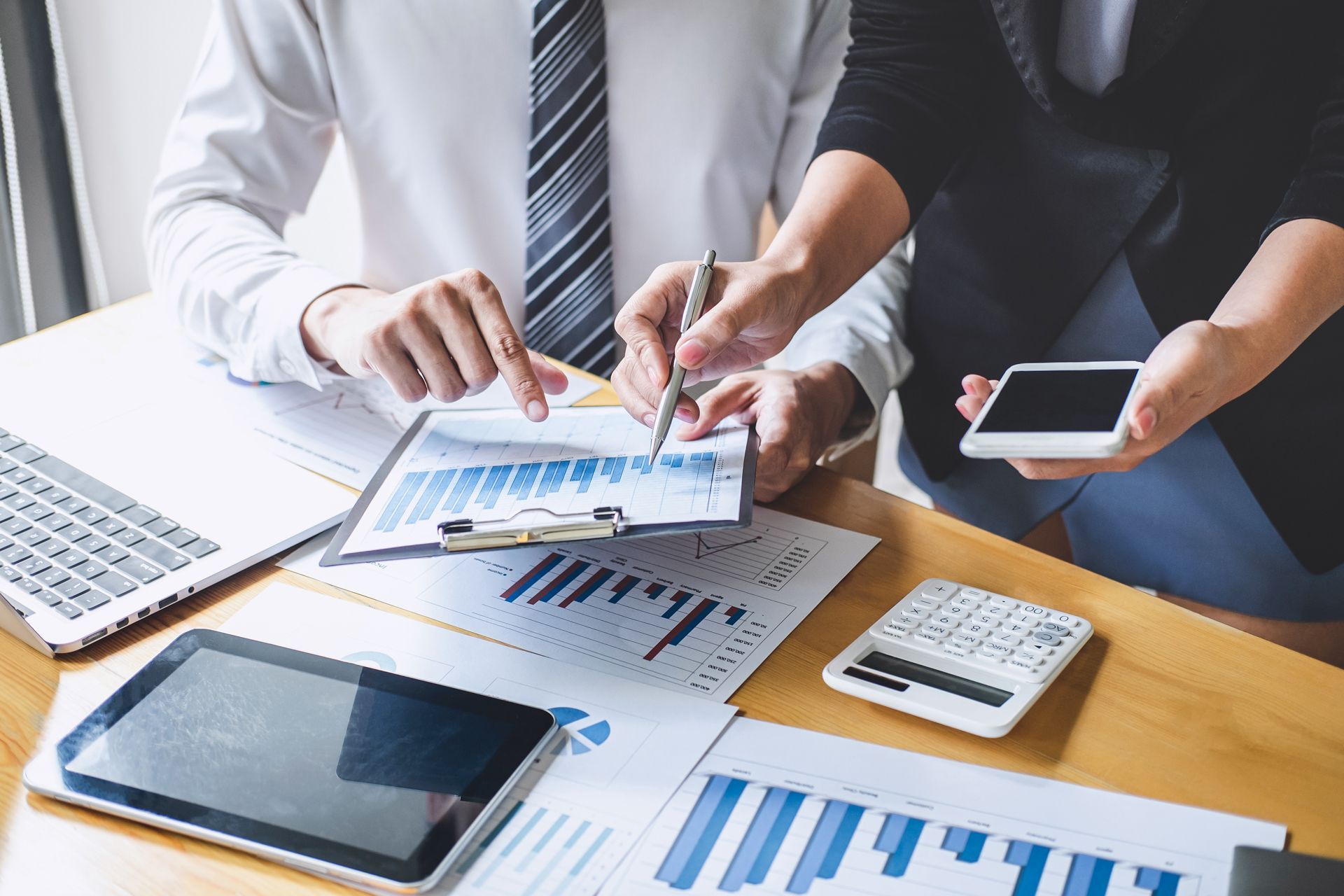 Two people reviewing financial charts and data on a desk with a laptop, tablet, calculator, and phone.