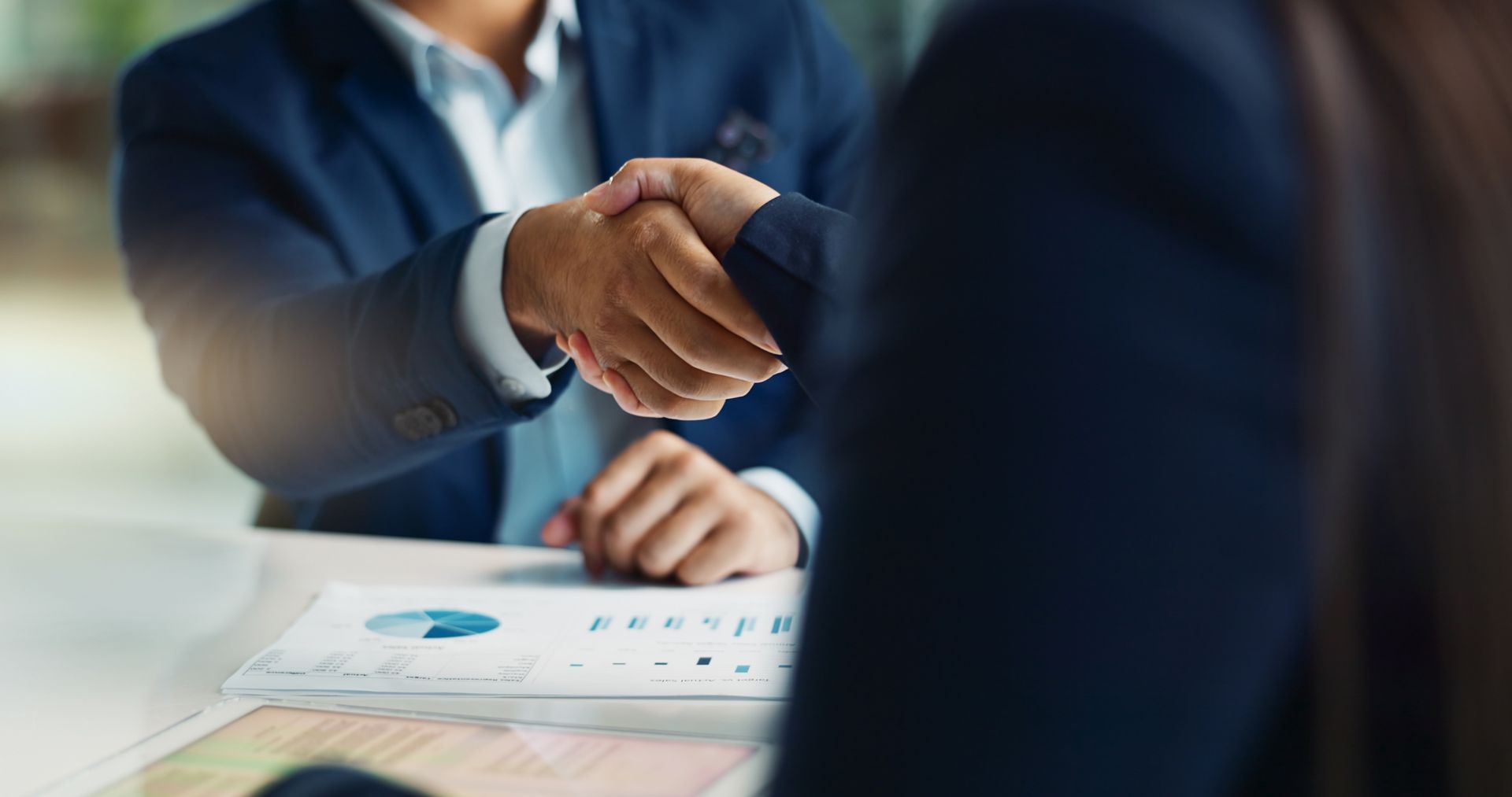 Two people in business suits shaking hands over a table with a chart.
