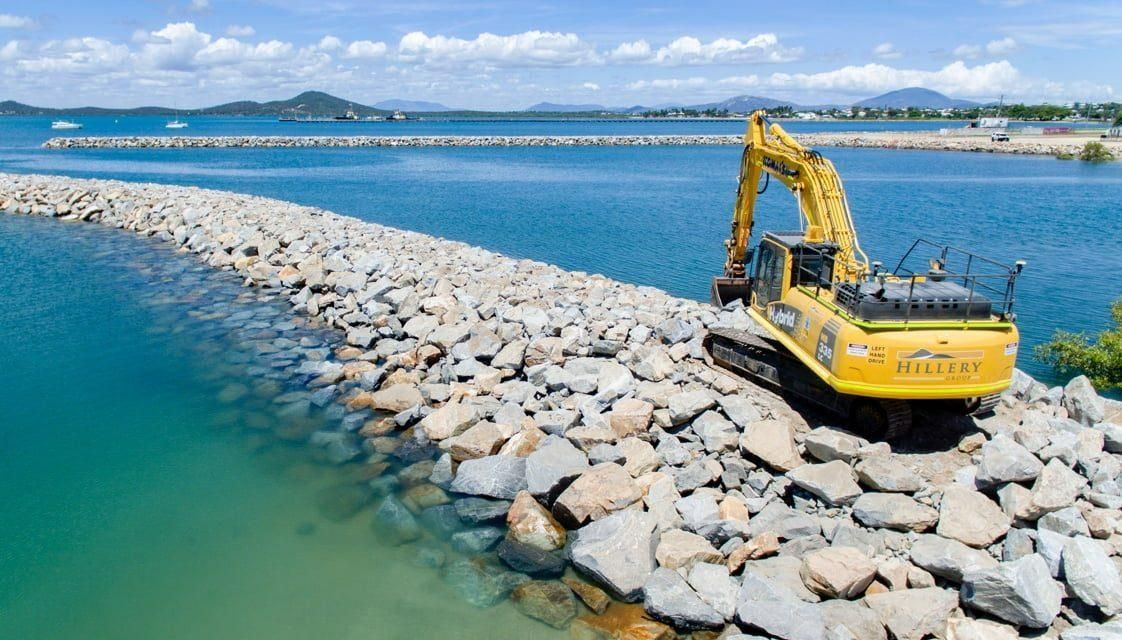 A Yellow Excavator Is Sitting on Top of A Pile of Rocks Next to A Body of Water — Hillery Group in Belmore, NSW